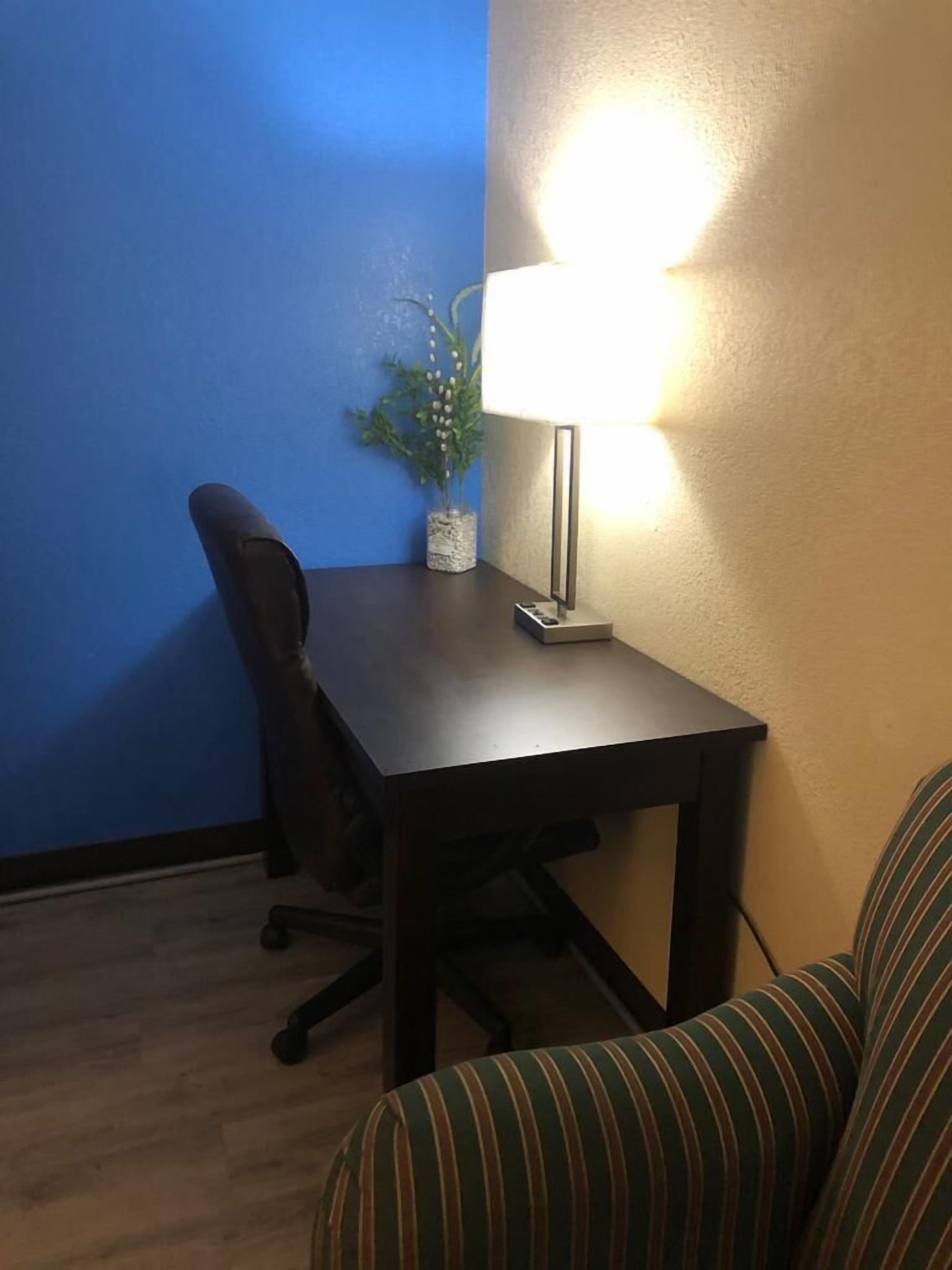 A small, dark wooden desk with a lamp, a chair, and a potted plant against a blue wall.