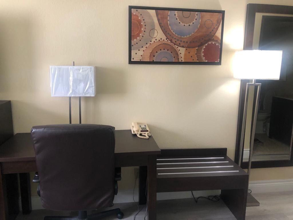 A hotel room with a desk, chair, lamp, and a painting on the wall.