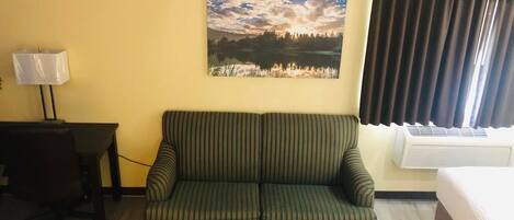 A room with a striped sofa, a small table with a lamp, a bed, and a painting of a landscape on the wall.