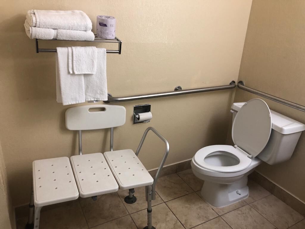 Une salle de bain équipée de toilettes, d’une barre d’appui, d’une étagère avec des serviettes et d’une petite table avec un tabouret.