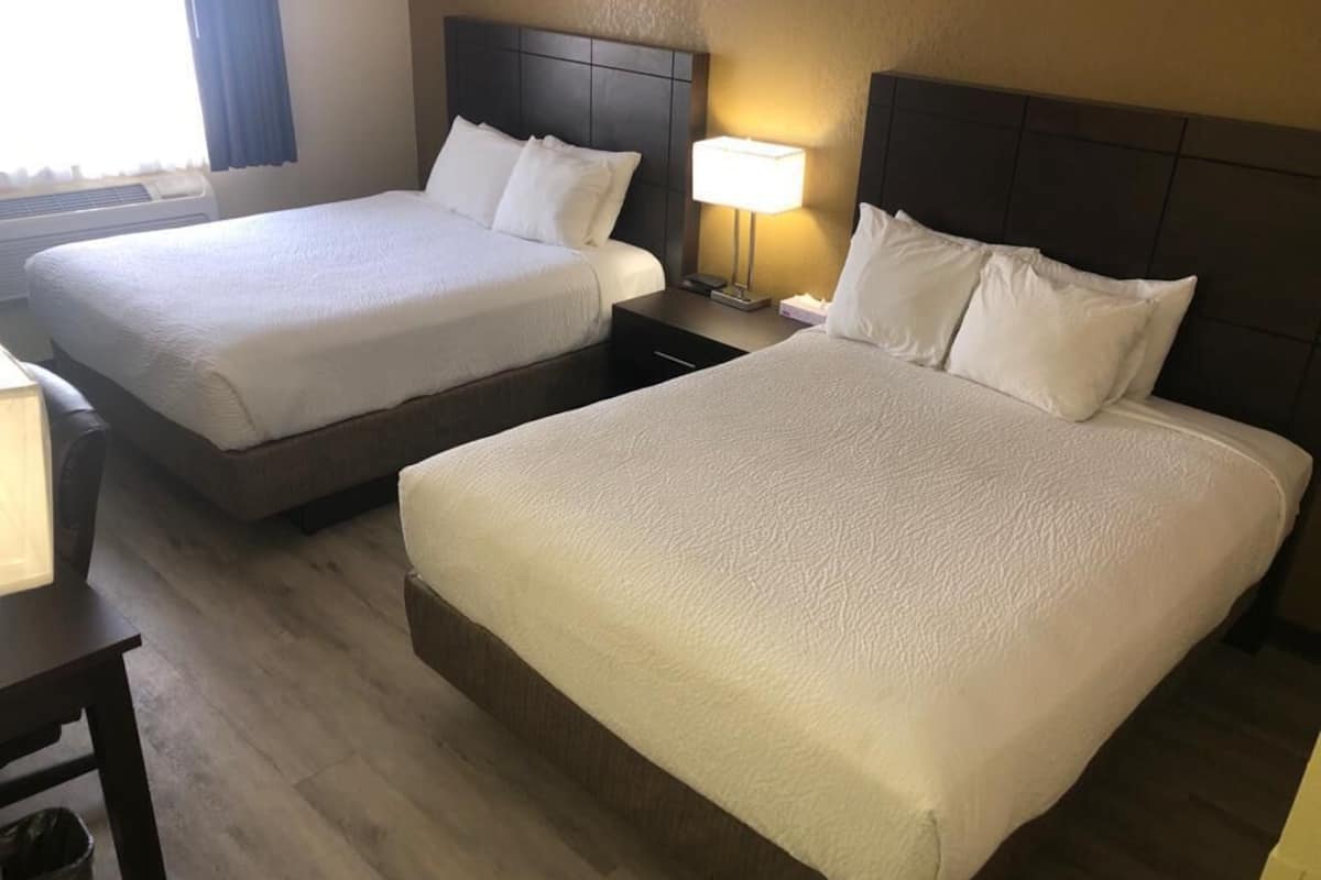 A hotel room with two beds, a bedside table, a lamp, and a window with curtains.