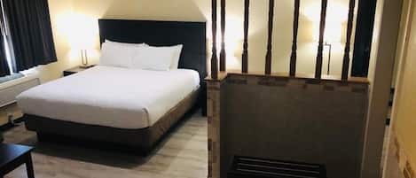 A hotel room with a large bed, a desk, a chair, a fireplace, and a mirror.