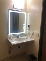 Casa de banho com lavatório branco, espelho com iluminação embutida, secador de cabelo e um cesto de lixo.