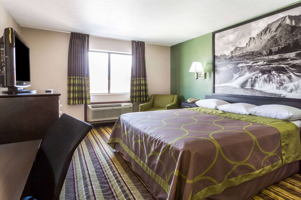 Hotelkamer met een bed, een bureau met een computer, een stoel, een televisie en een groot raam met gordijnen.