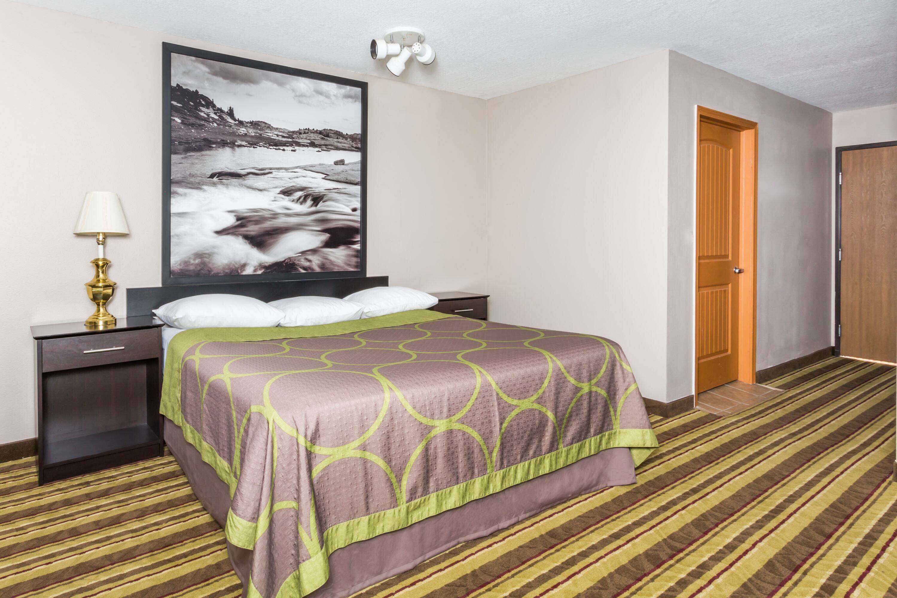A hotel room with a bed, a nightstand, a lamp, a painting, and a door.