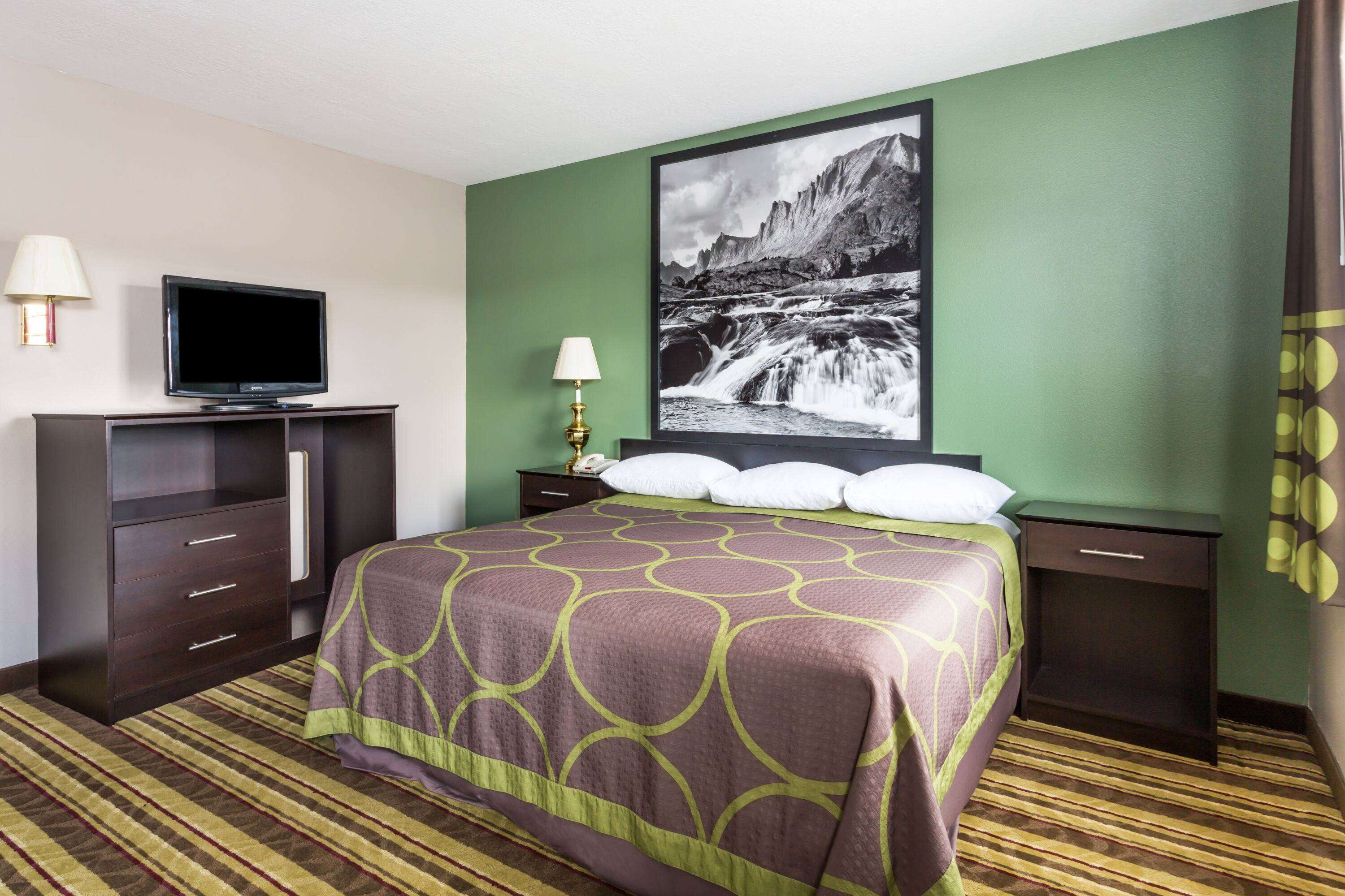 A hotel room with a bed, a television, a nightstand, a lamp, and a painting on the wall.