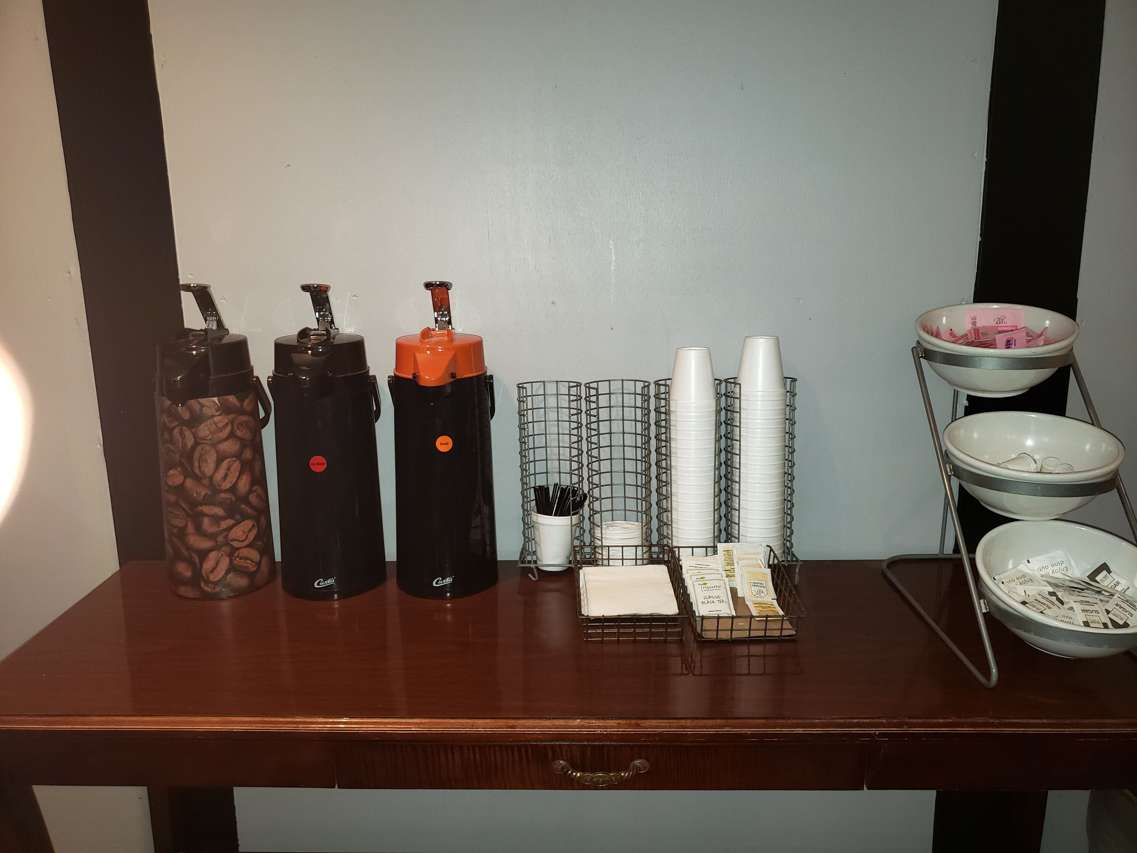 A wooden table with coffee dispensers, a wire basket with napkins, and a tray with bowls and packets.
