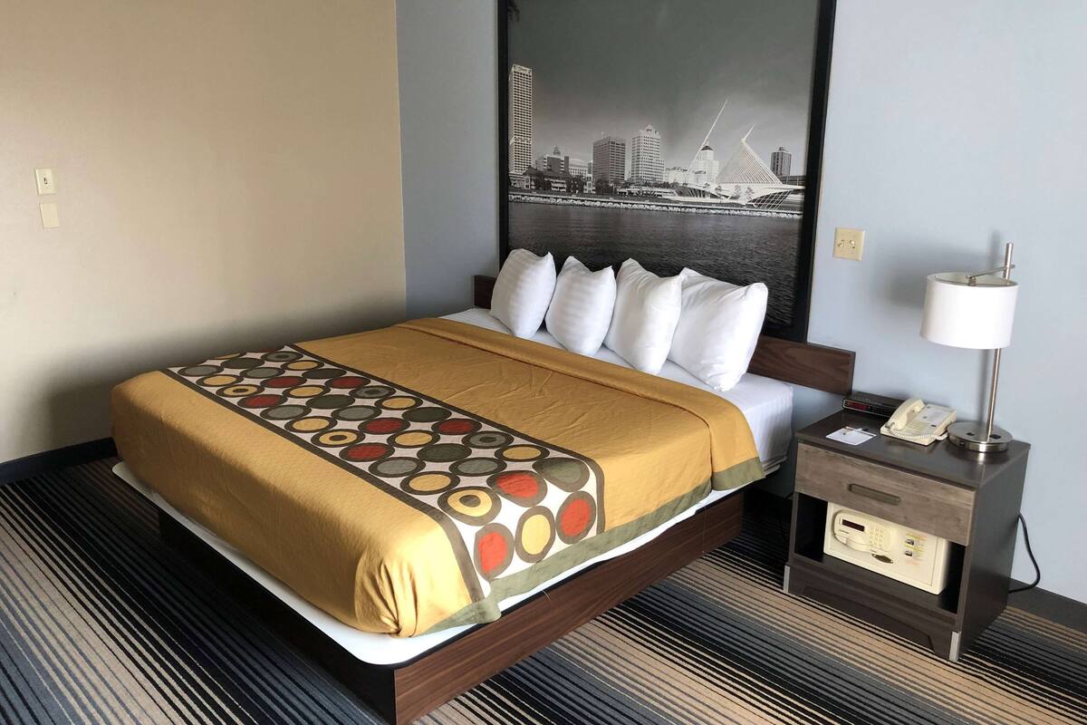 A hotel room with a bed, a nightstand, a lamp, and a cityscape painting on the wall.