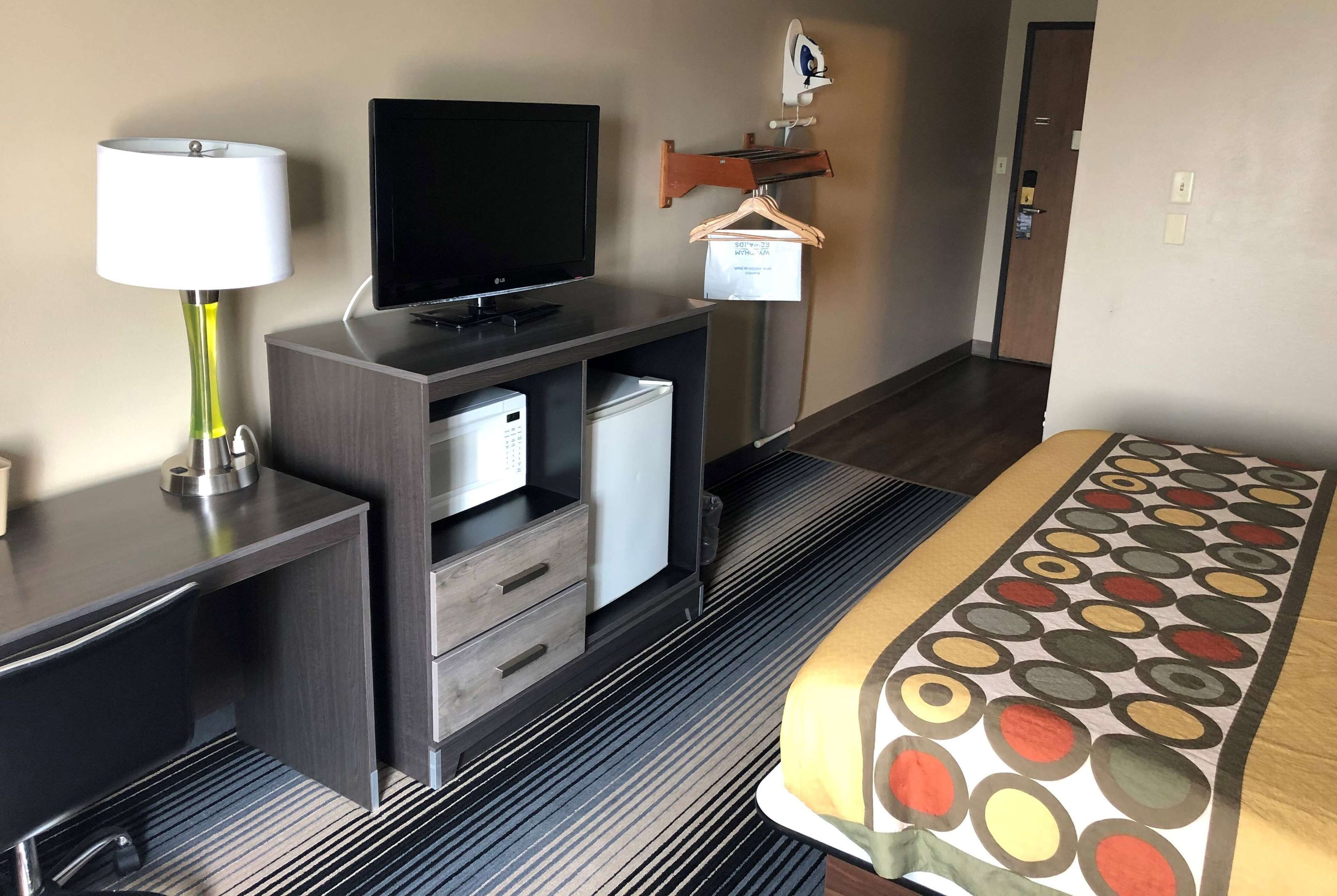 Quarto de hotel com televisão, luminária, mini-geladeira, cama com colcha estampada e cabideiro fixado na parede.