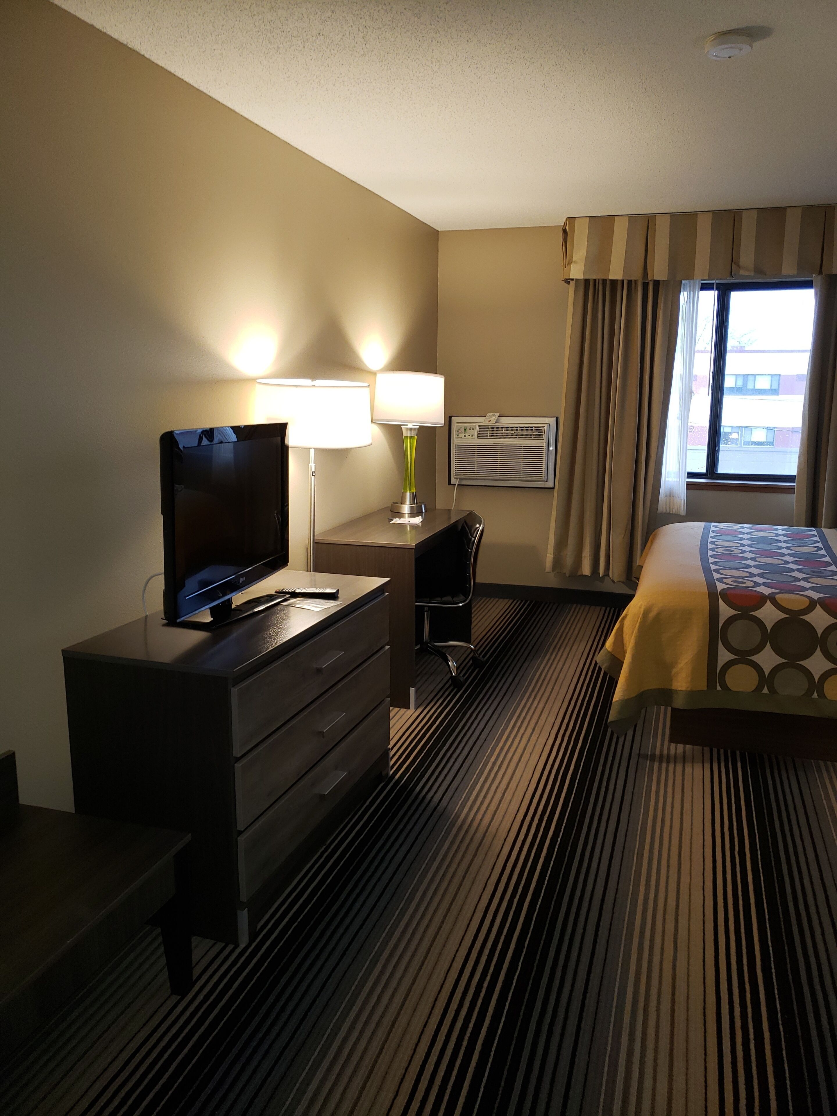 A hotel room with a bed, a desk with a computer, a TV, a lamp, and a window with curtains.