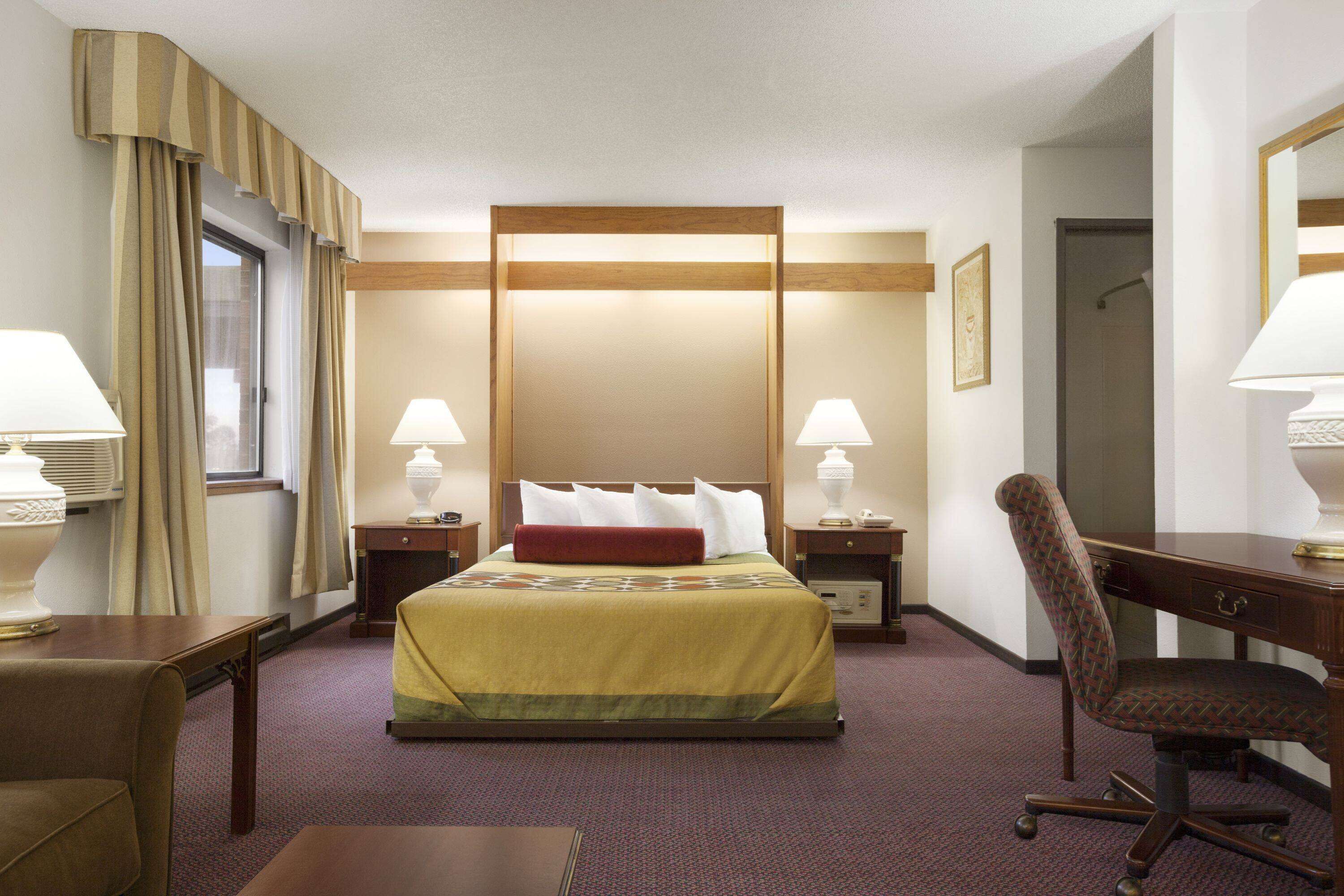 Una habitación de hotel con una cama grande, dos mesitas de noche con lámparas, un escritorio con silla y un ventanal con cortinas.