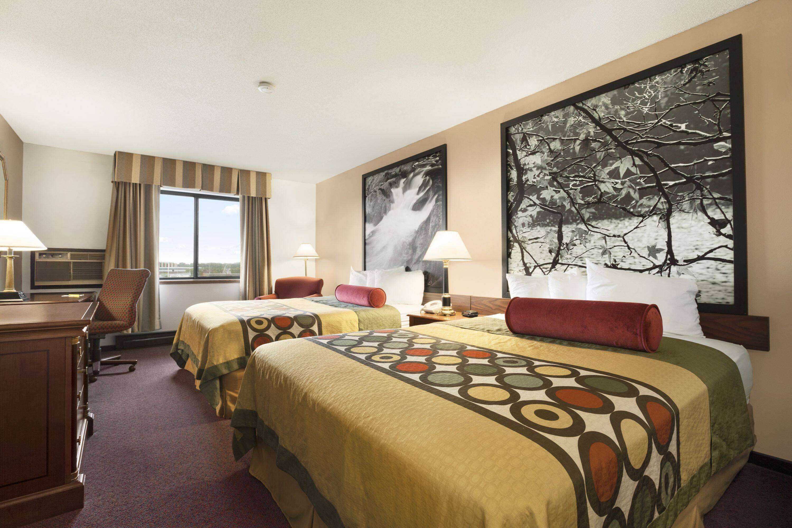 A hotel room with two beds, a desk, and a chair. There is a large window with curtains, a painting on the wall, and a lamp on the desk.