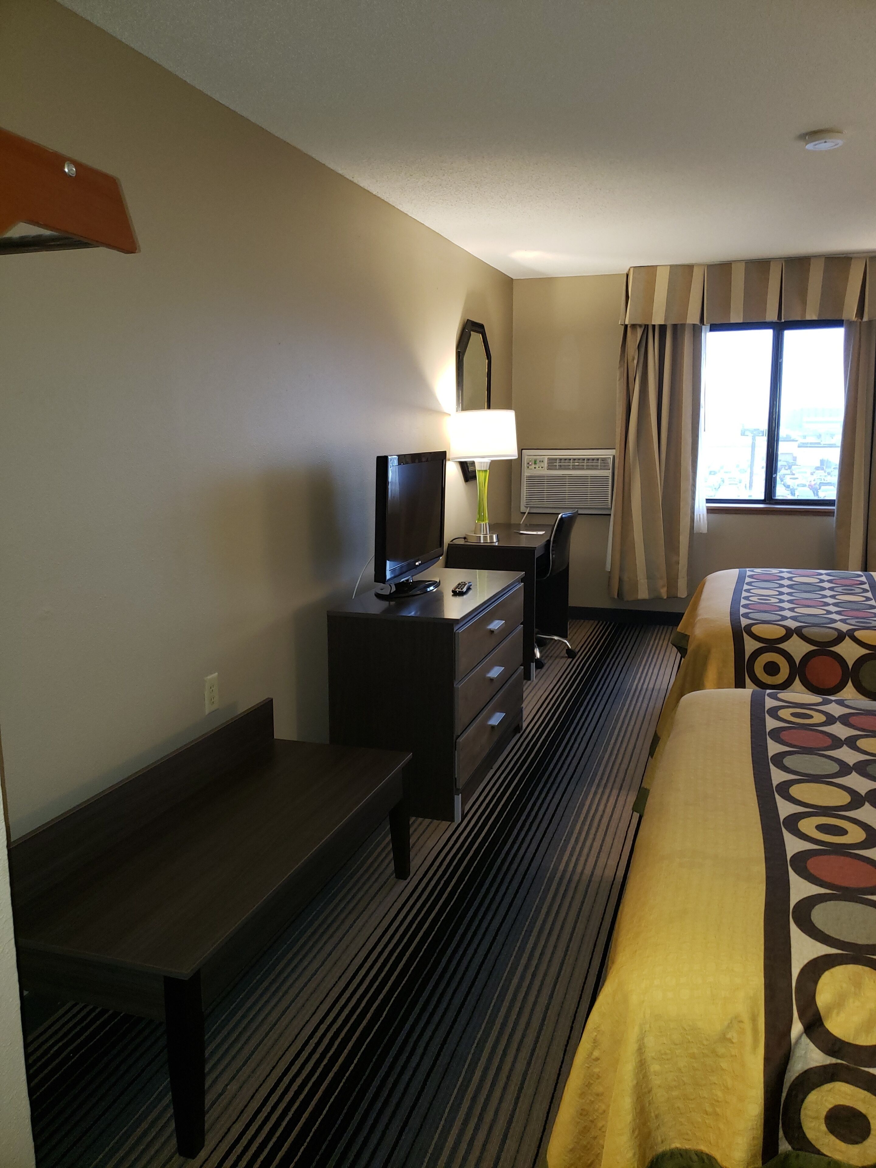 Une chambre d’hôtel avec un lit, un bureau, une télévision, une lampe et une fenêtre avec des rideaux.
