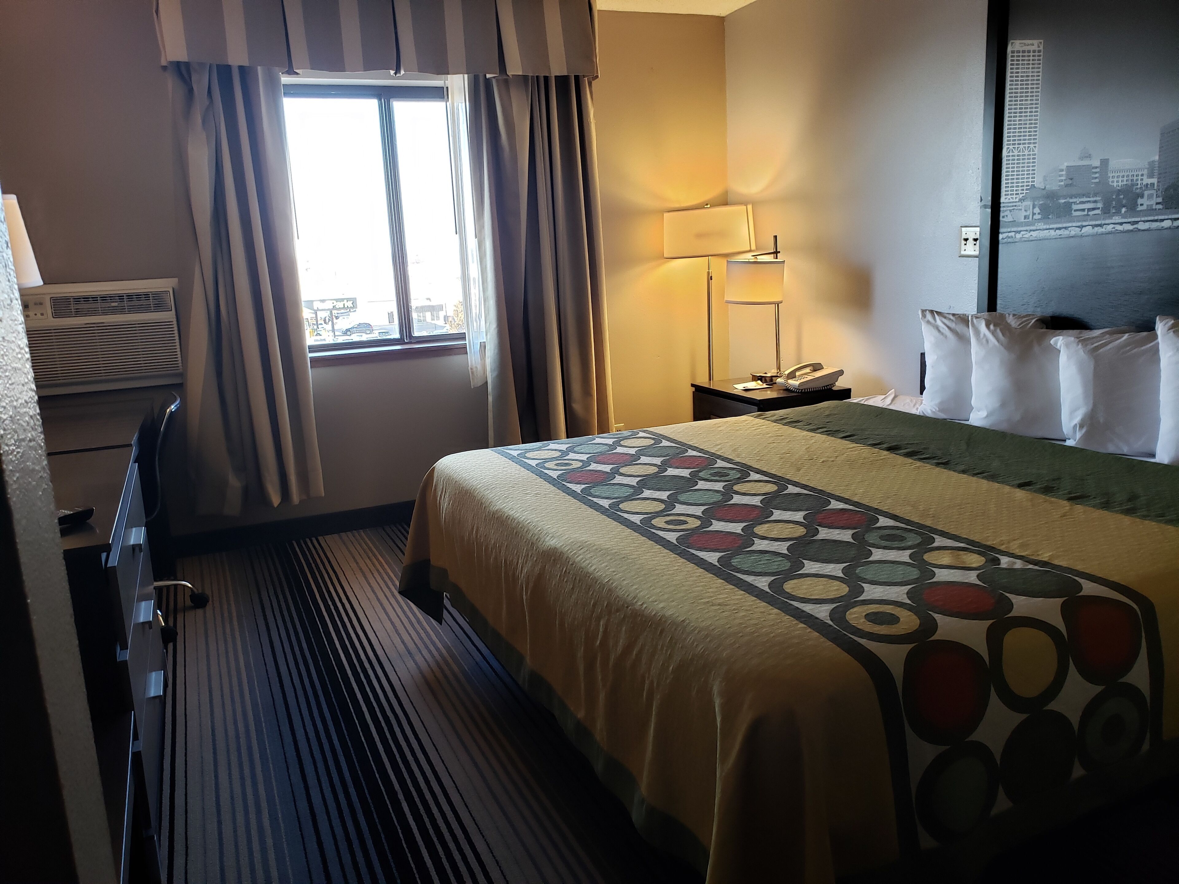 Una habitación de hotel con cama, lámpara de noche, teléfono y ventana con cortinas.