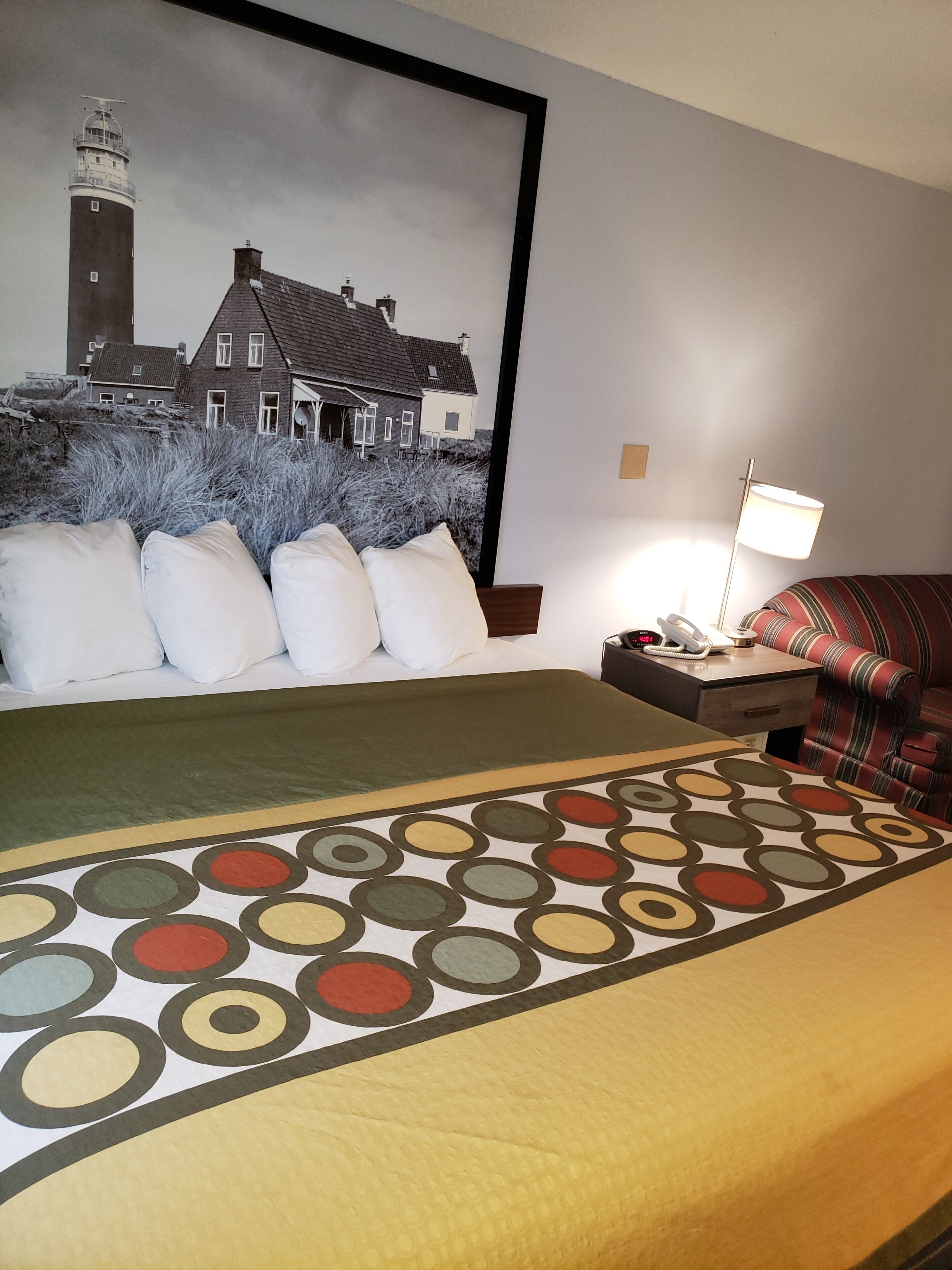 A hotel room with a large bed, a nightstand with a lamp, a striped sofa, and a wall painting of a lighthouse and house.