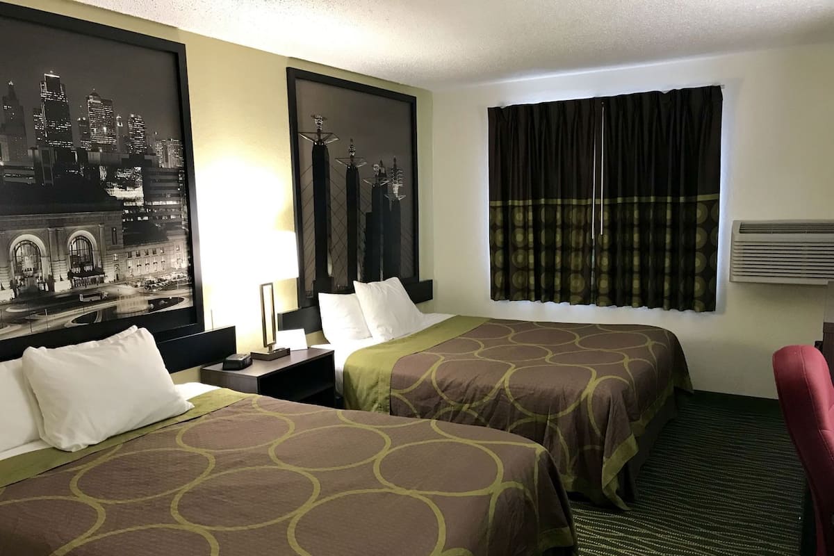 A hotel room with two beds, a large framed cityscape picture, a window with curtains, and a red chair.