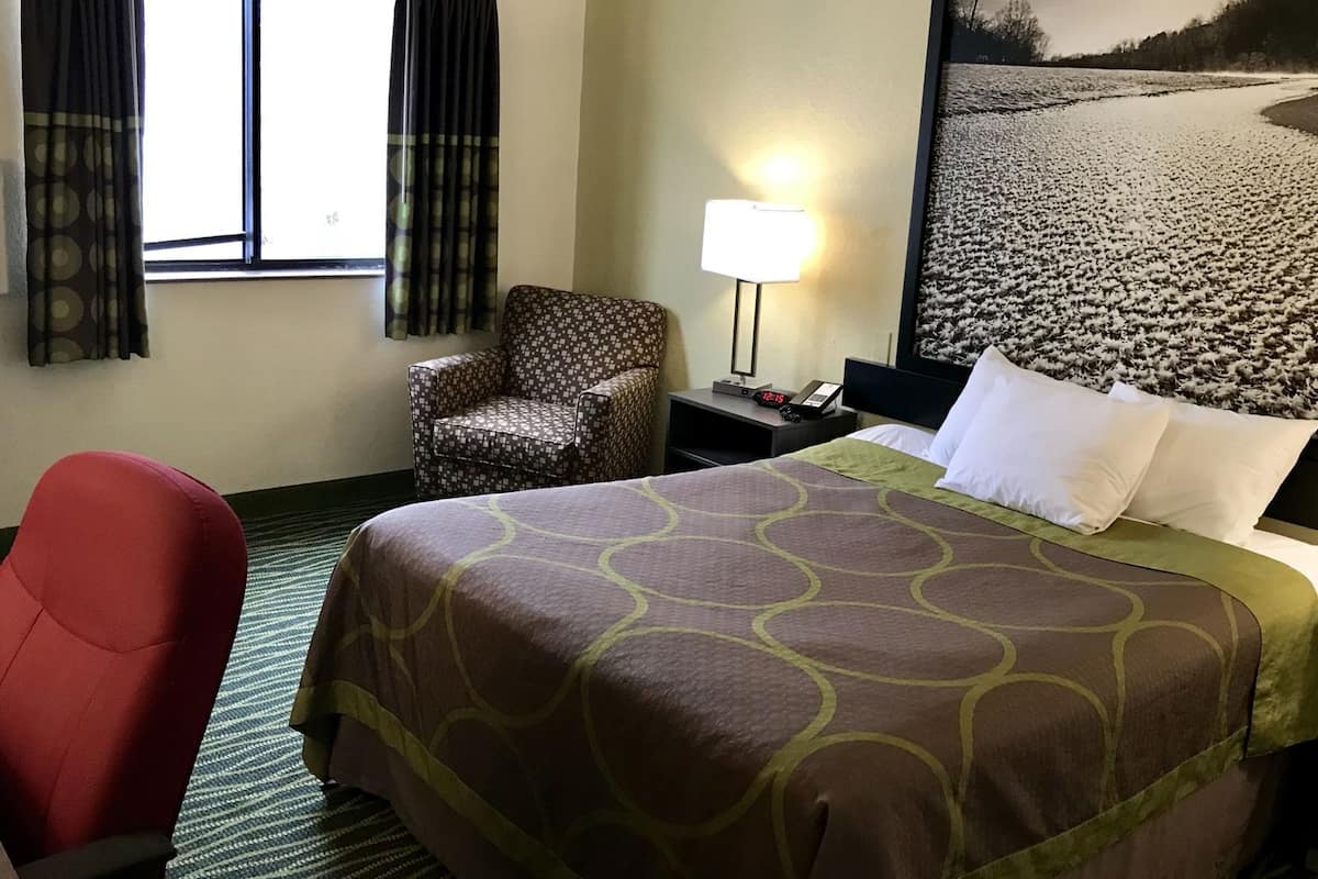 A hotel room with a bed, a chair, a nightstand with a lamp, and a window with curtains.