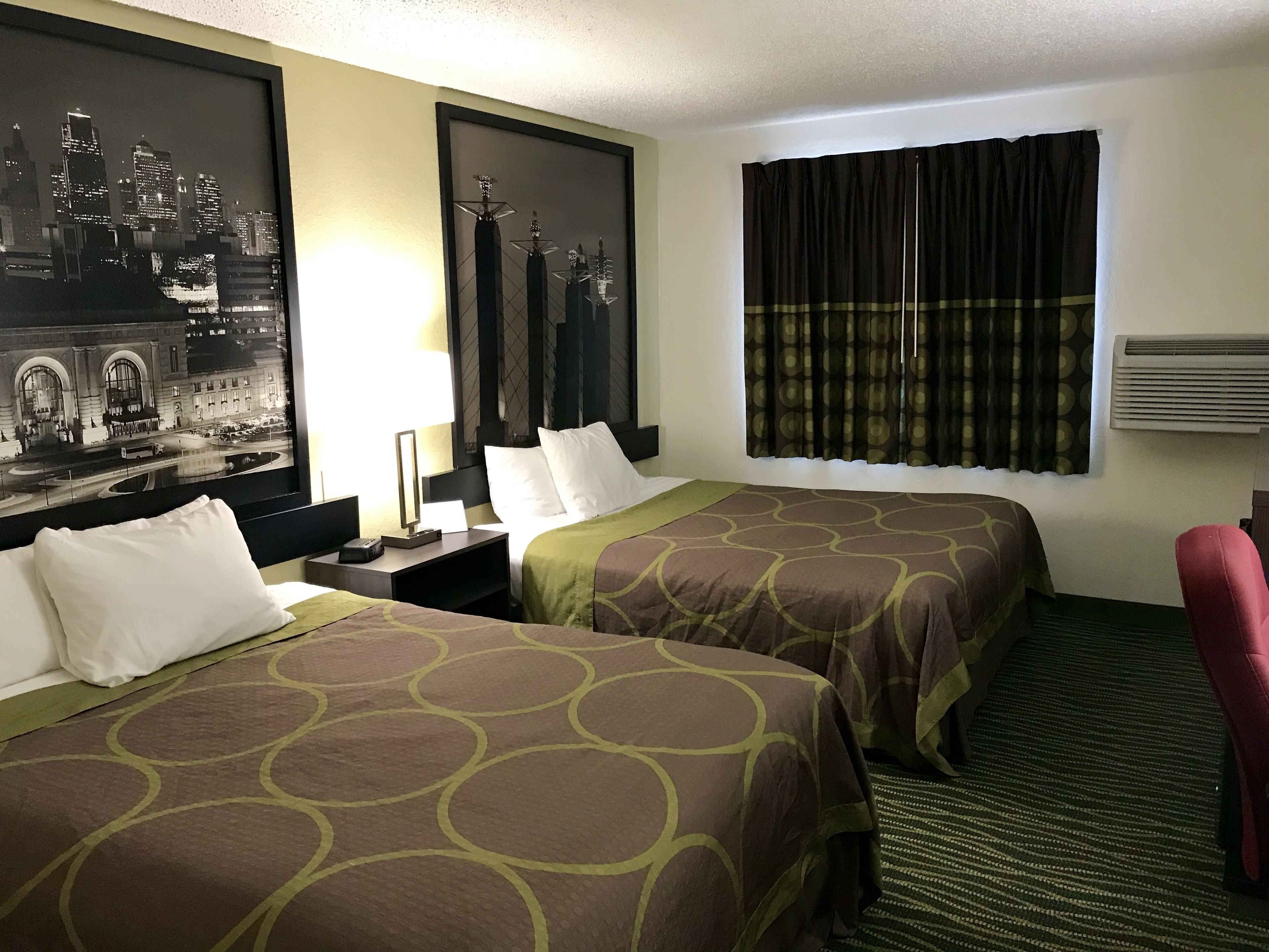 A hotel room with two beds, a large framed cityscape picture, a window with curtains, and a red chair.