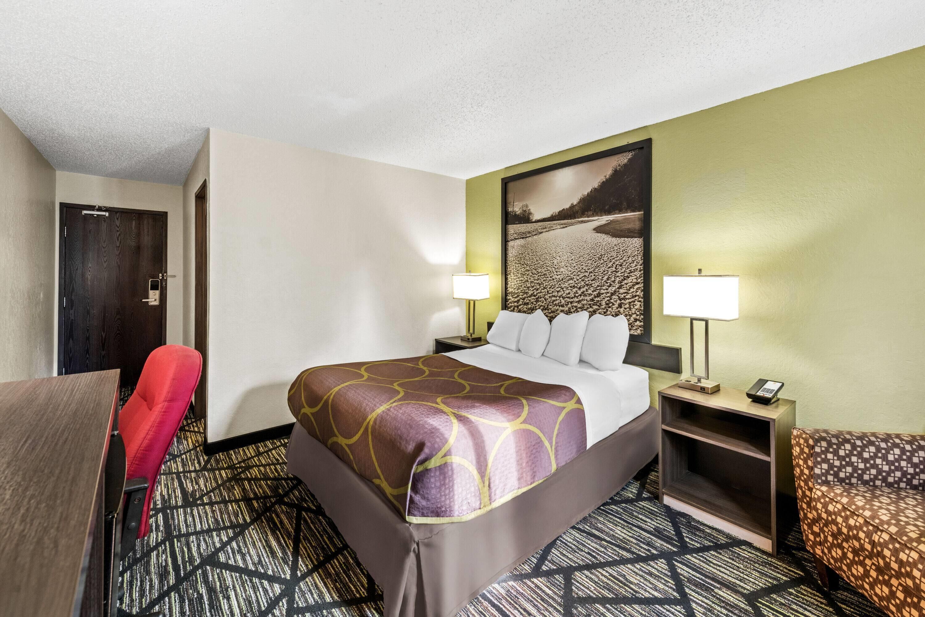 A hotel room with a bed, two bedside lamps, a red chair, a brown sofa, and a large wall painting.