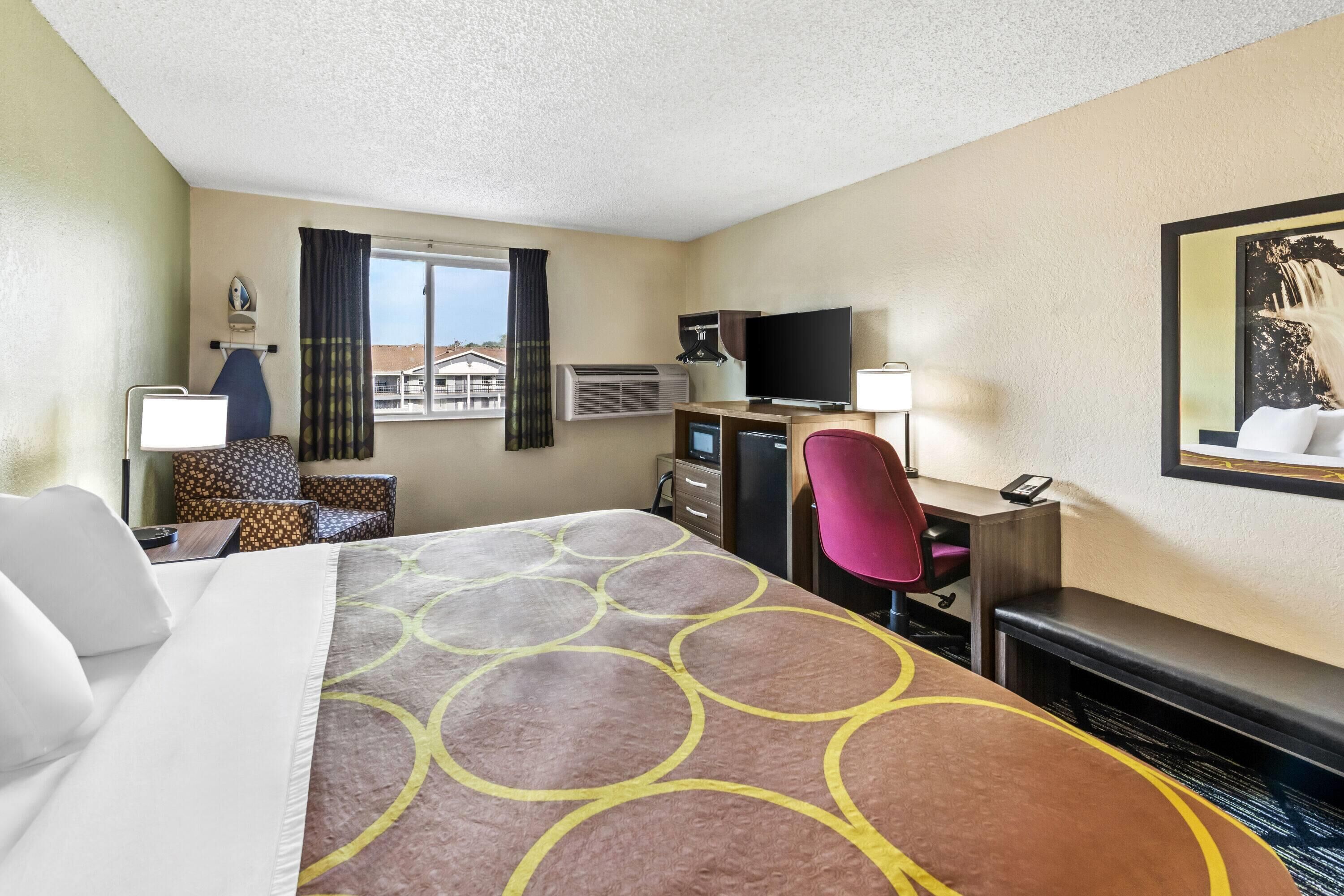 Une chambre d’hôtel avec un lit, un bureau avec un ordinateur, une télévision et une fenêtre avec des rideaux.