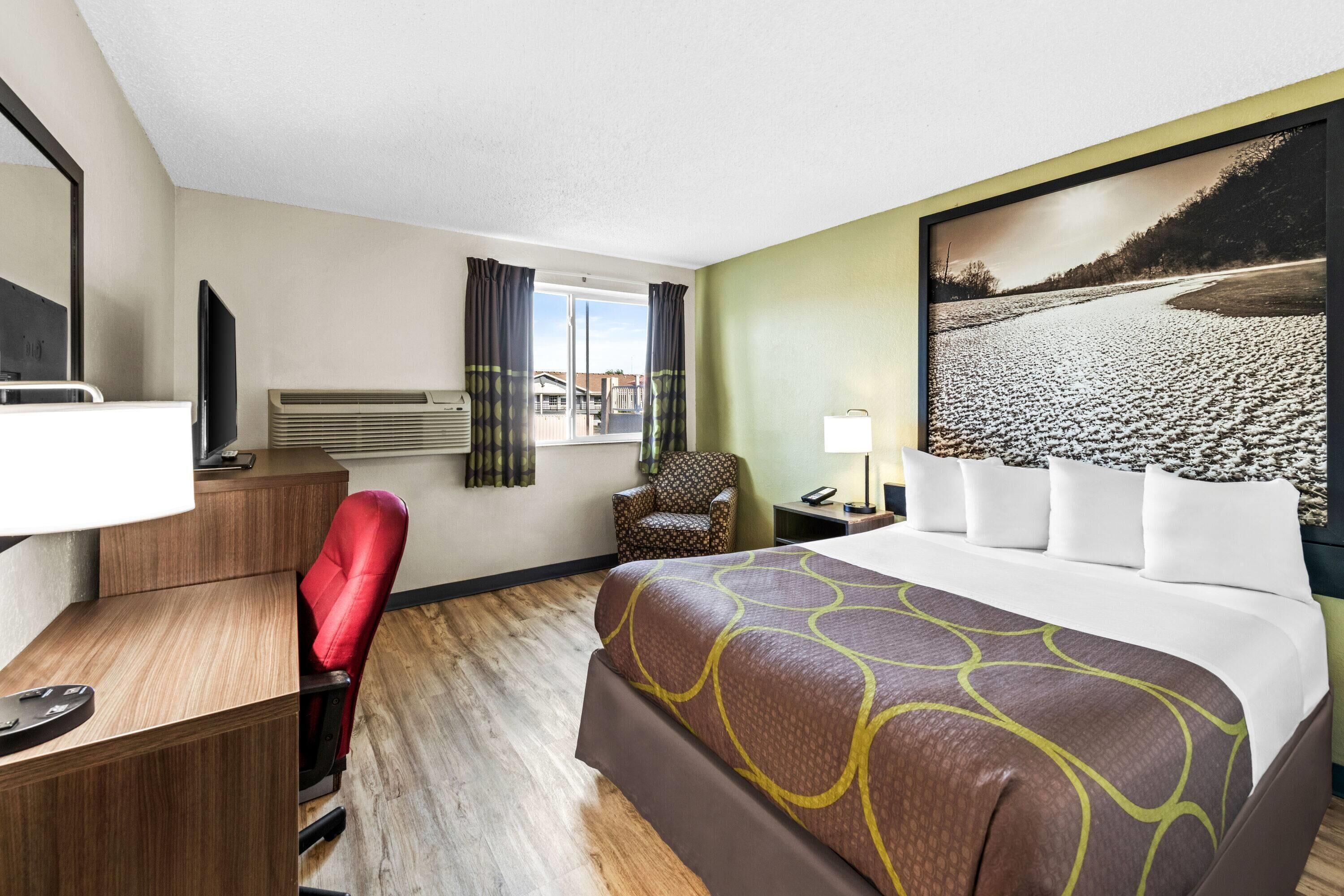 A hotel room with a large bed, a desk with a chair, a TV, an air conditioning unit, a chair, a nightstand, a lamp, and a window with curtains.
