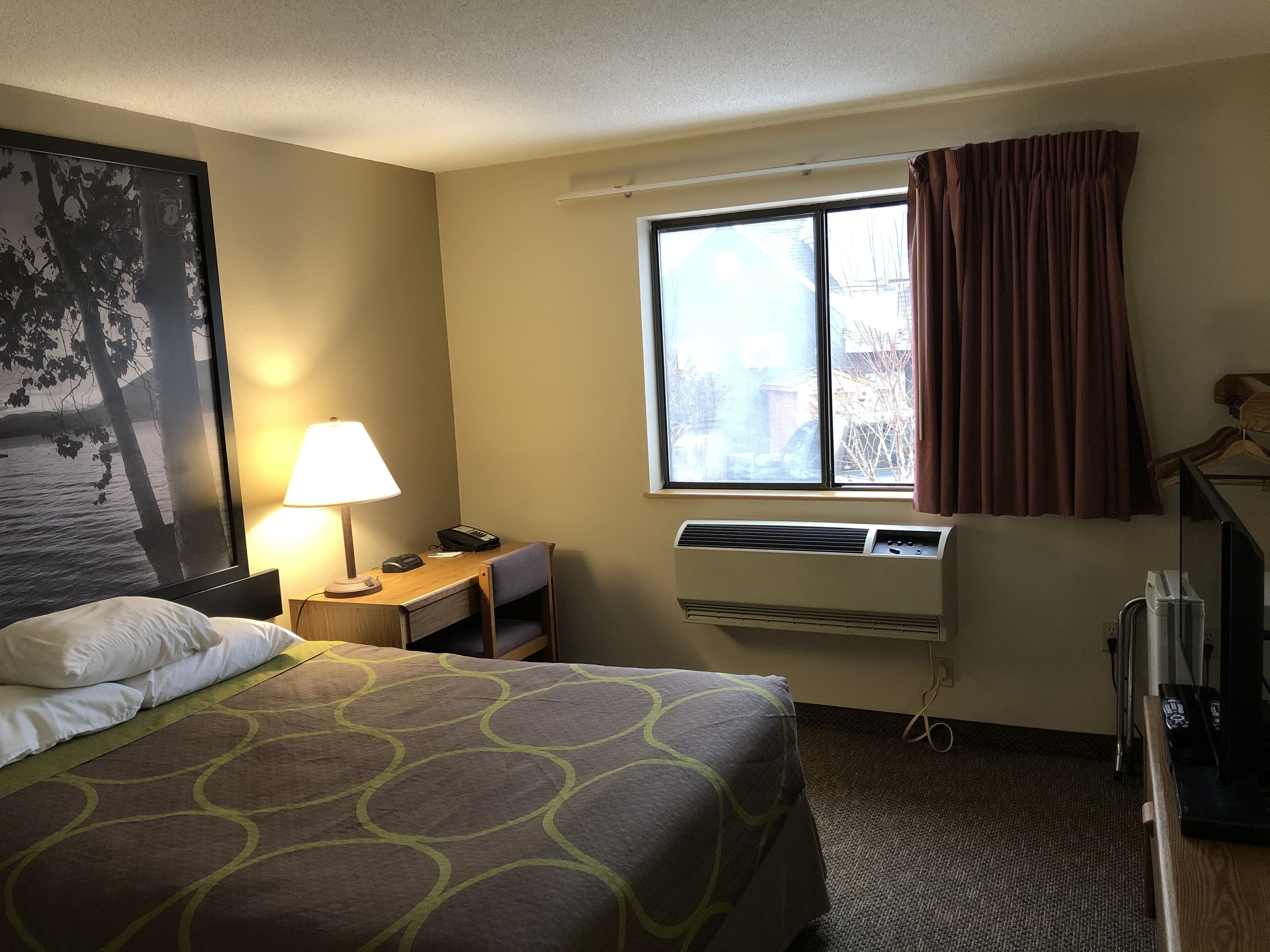 A hotel room with a bed, a desk, a lamp, an air conditioner, and a window with curtains.