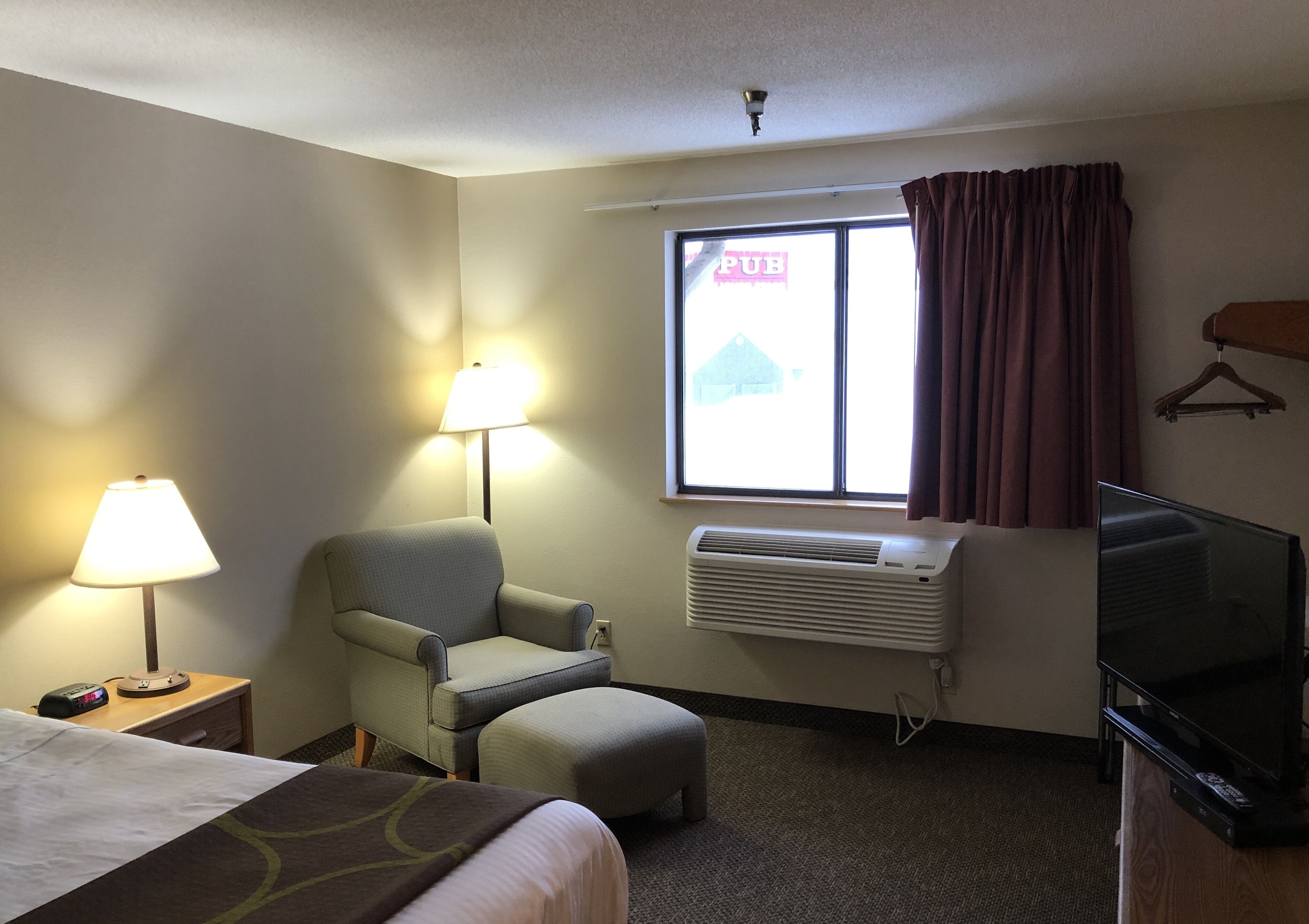 A hotel room with a bed, a chair, a TV, a lamp, an air conditioner, and a window with a view of a building labeled "PUB".