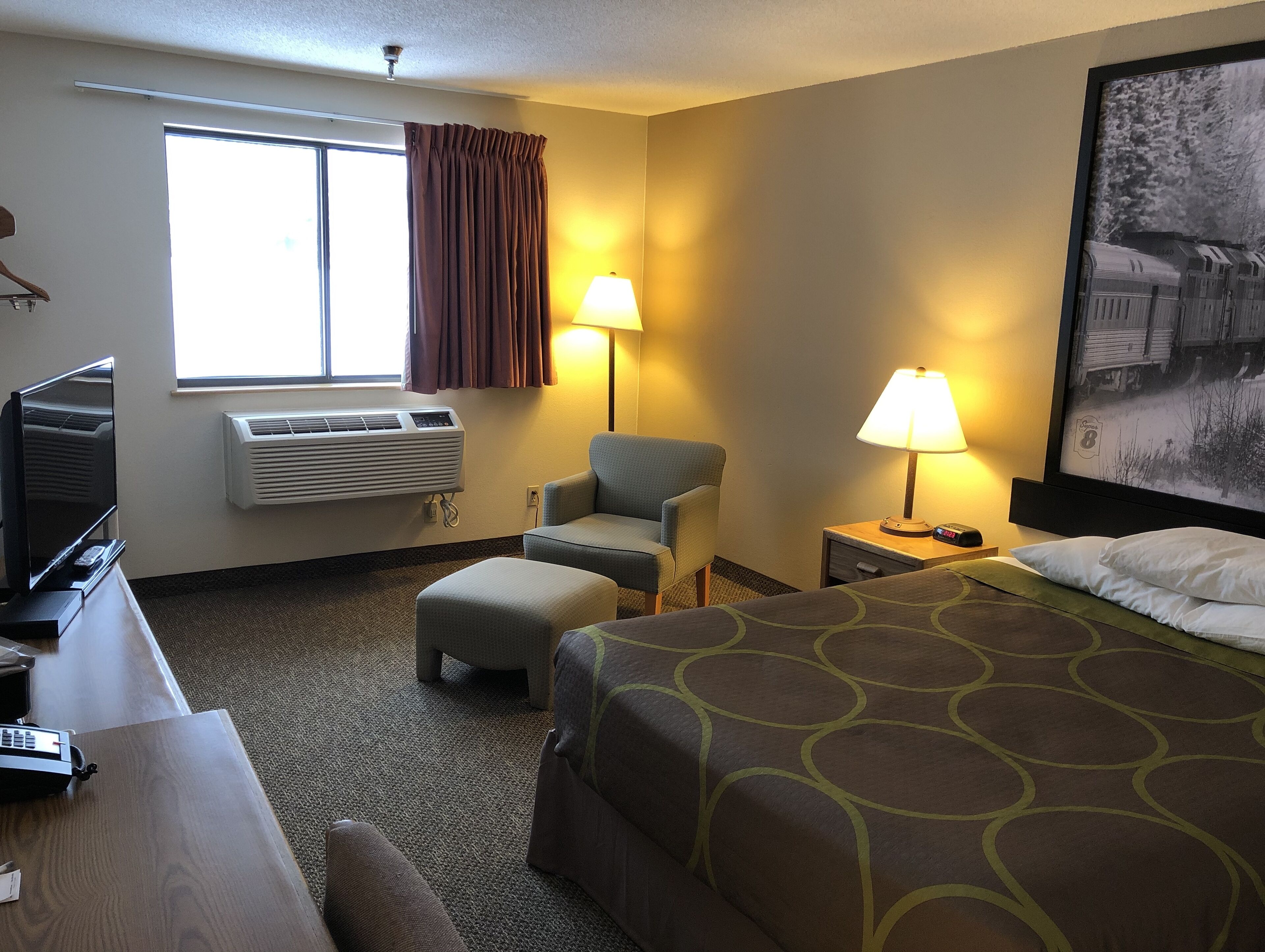 A hotel room with a bed, a desk with a television, a chair, two bedside lamps, and a large window with curtains.