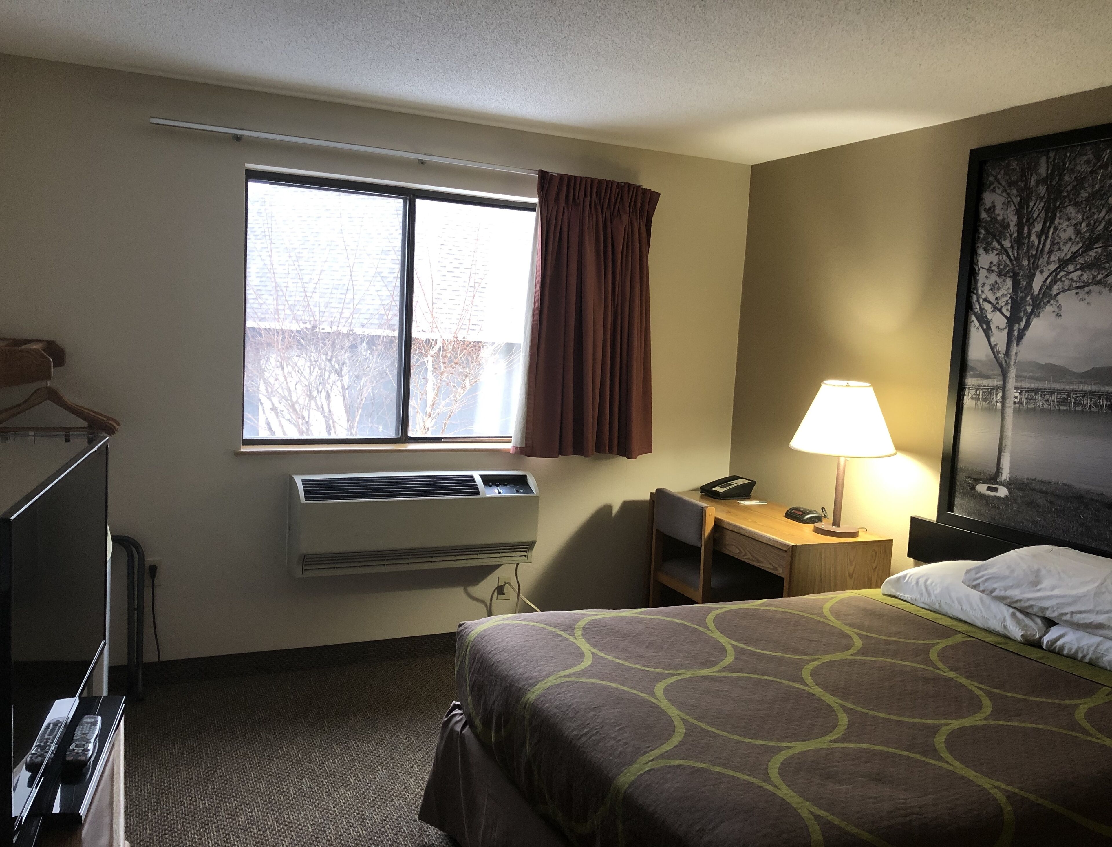 Quarto de hotel com cama, escrivaninha com luminária, janela com cortinas, ar condicionado e um quadro na parede.