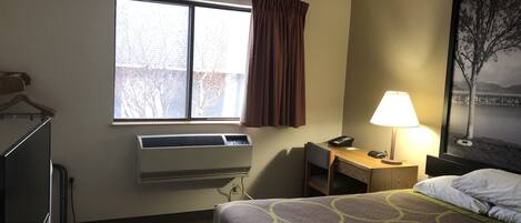 Quarto de hotel com cama, escrivaninha com luminária, janela com cortinas, ar condicionado e um quadro na parede.