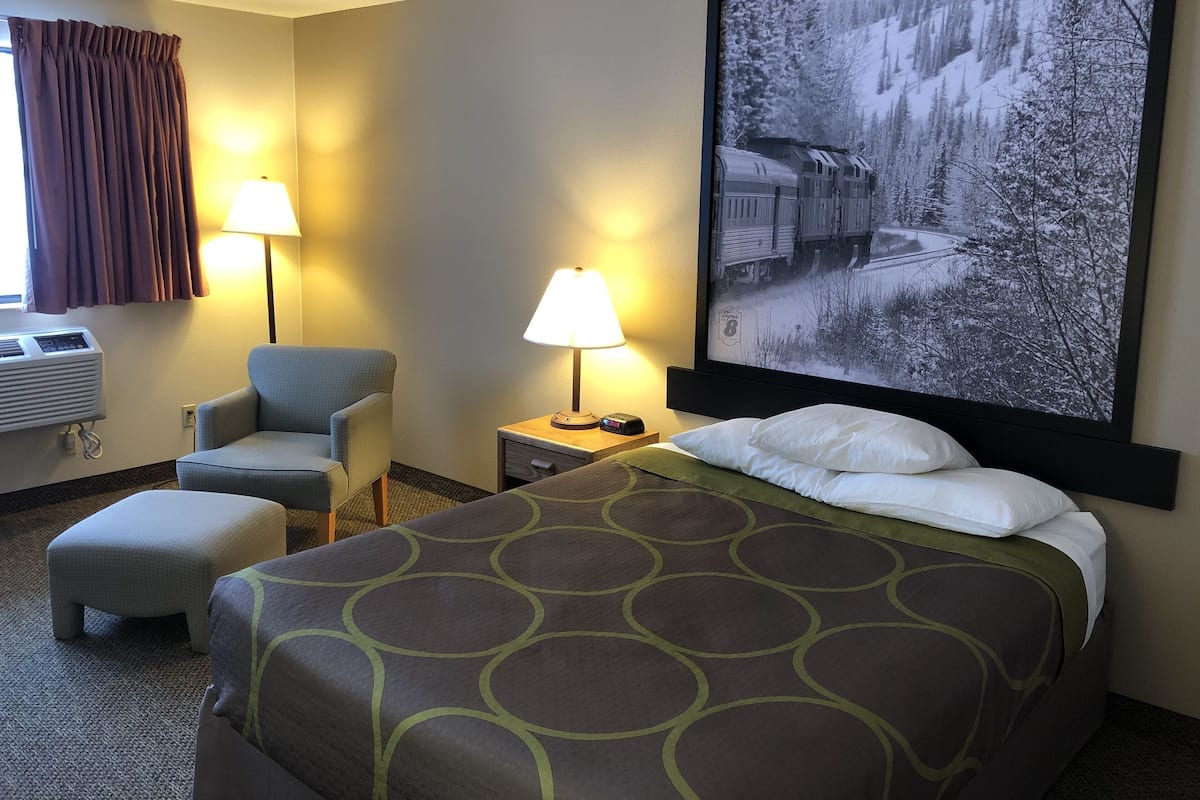 Habitación de hotel con cama, dos lámparas de noche, una silla, aire acondicionado y un cuadro grande en la pared.