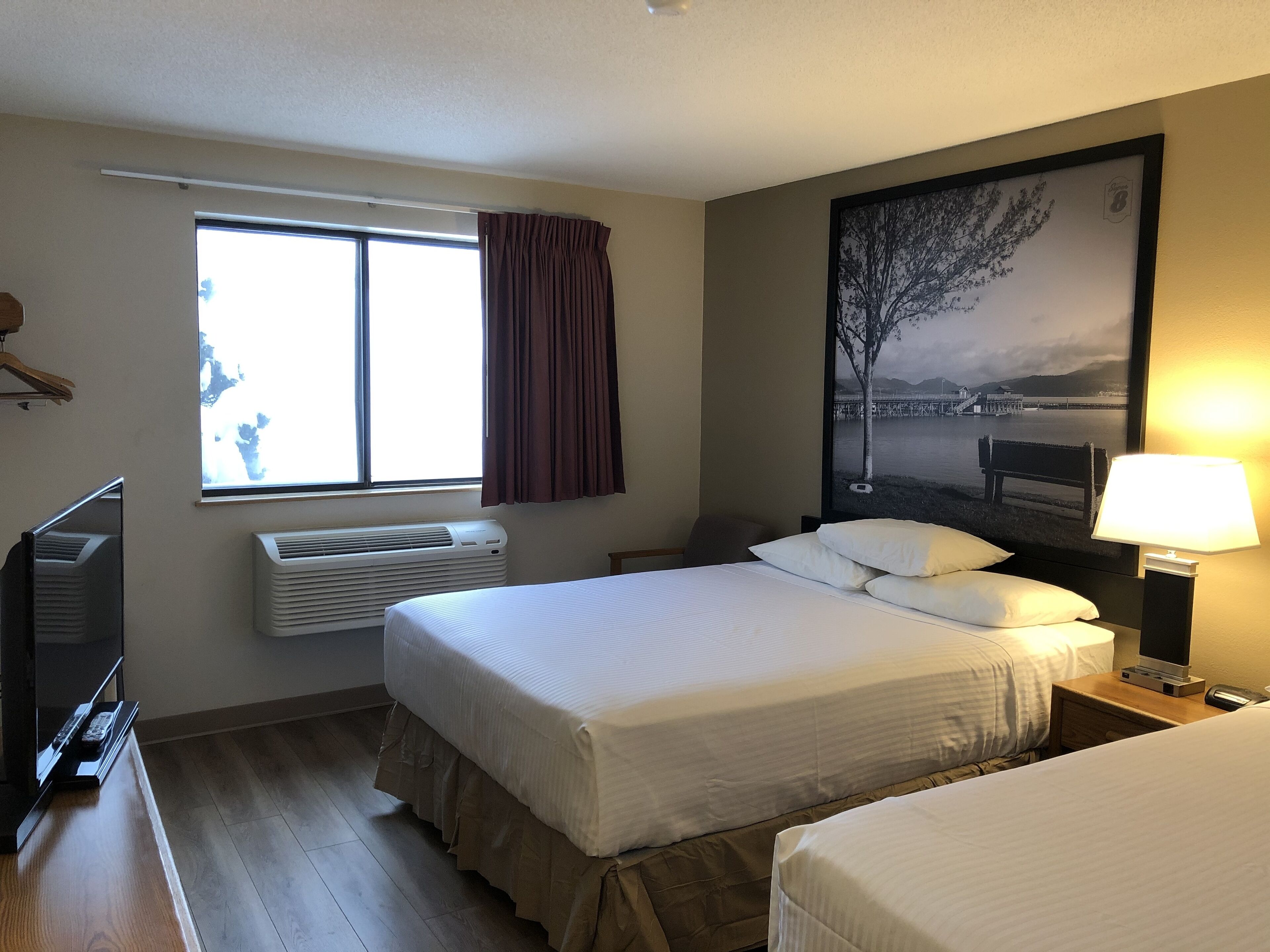 A hotel room with a bed, a TV, an air conditioner, a window with curtains, and a painting on the wall.