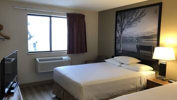 A hotel room with a bed, a TV, an air conditioner, a window with curtains, and a painting on the wall.
