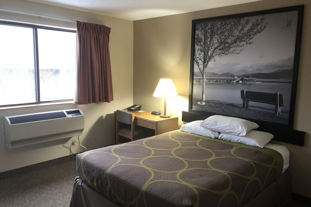 Habitación de hotel con cama, escritorio con lámpara, teléfono, ventana con cortinas e imagen enmarcada en la pared.