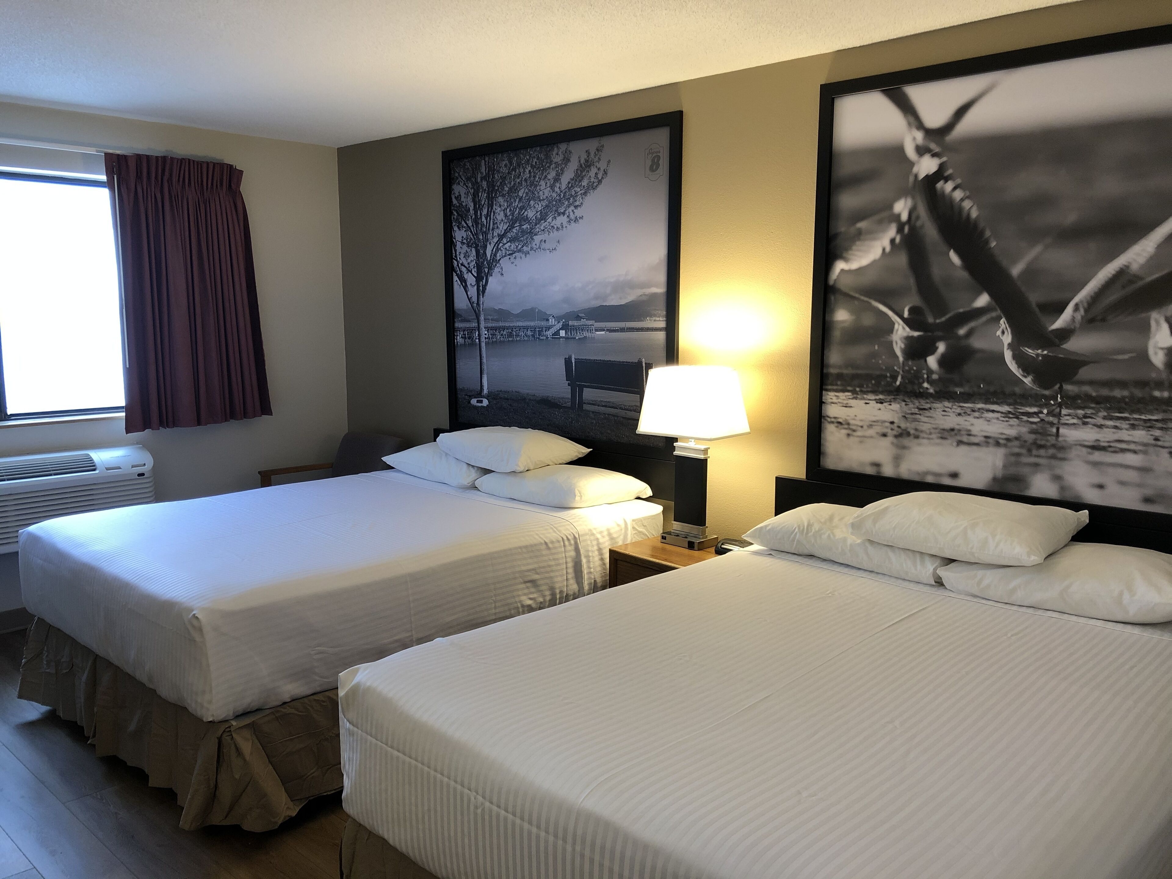 Een hotelkamer met twee bedden, een raam met gordijnen, een wandairconditioner en twee ingelijste foto's.
