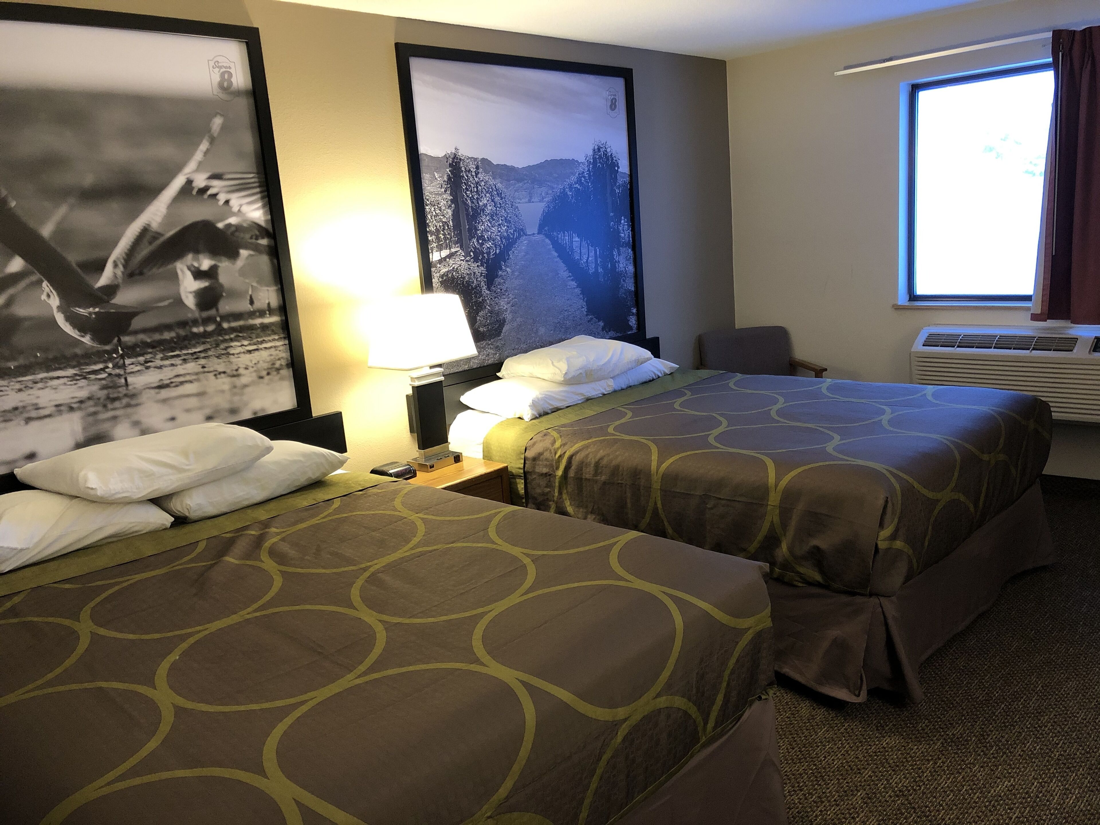 Een hotelkamer met twee bedden, een nachtkastje met een lamp, een stoel en een groot raam met gordijnen.