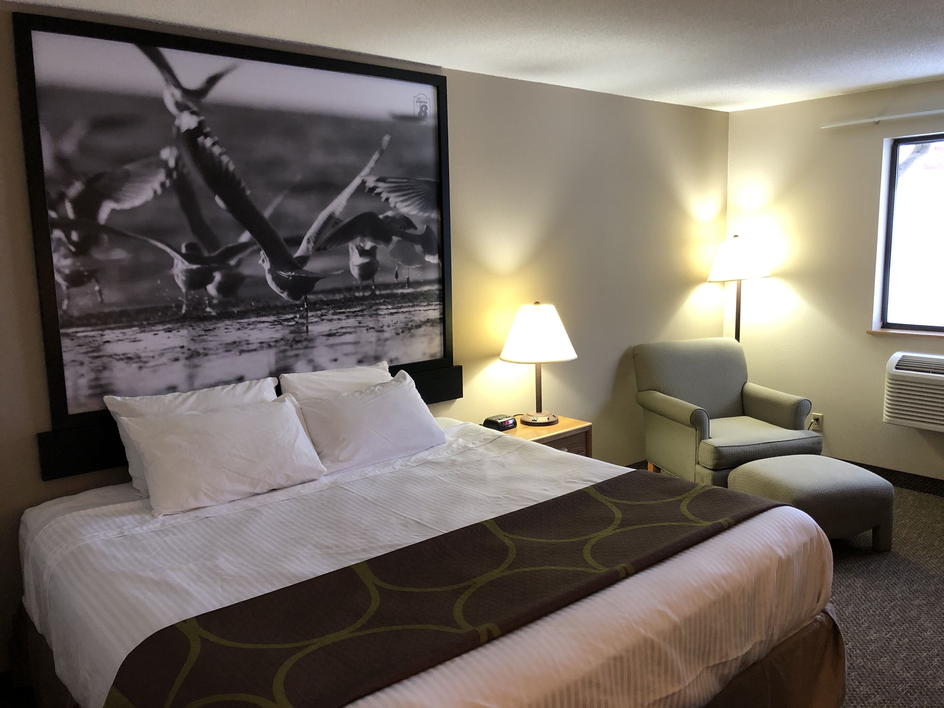 Habitación de hotel con cama, una silla, una lámpara y un cuadro grande en la pared.