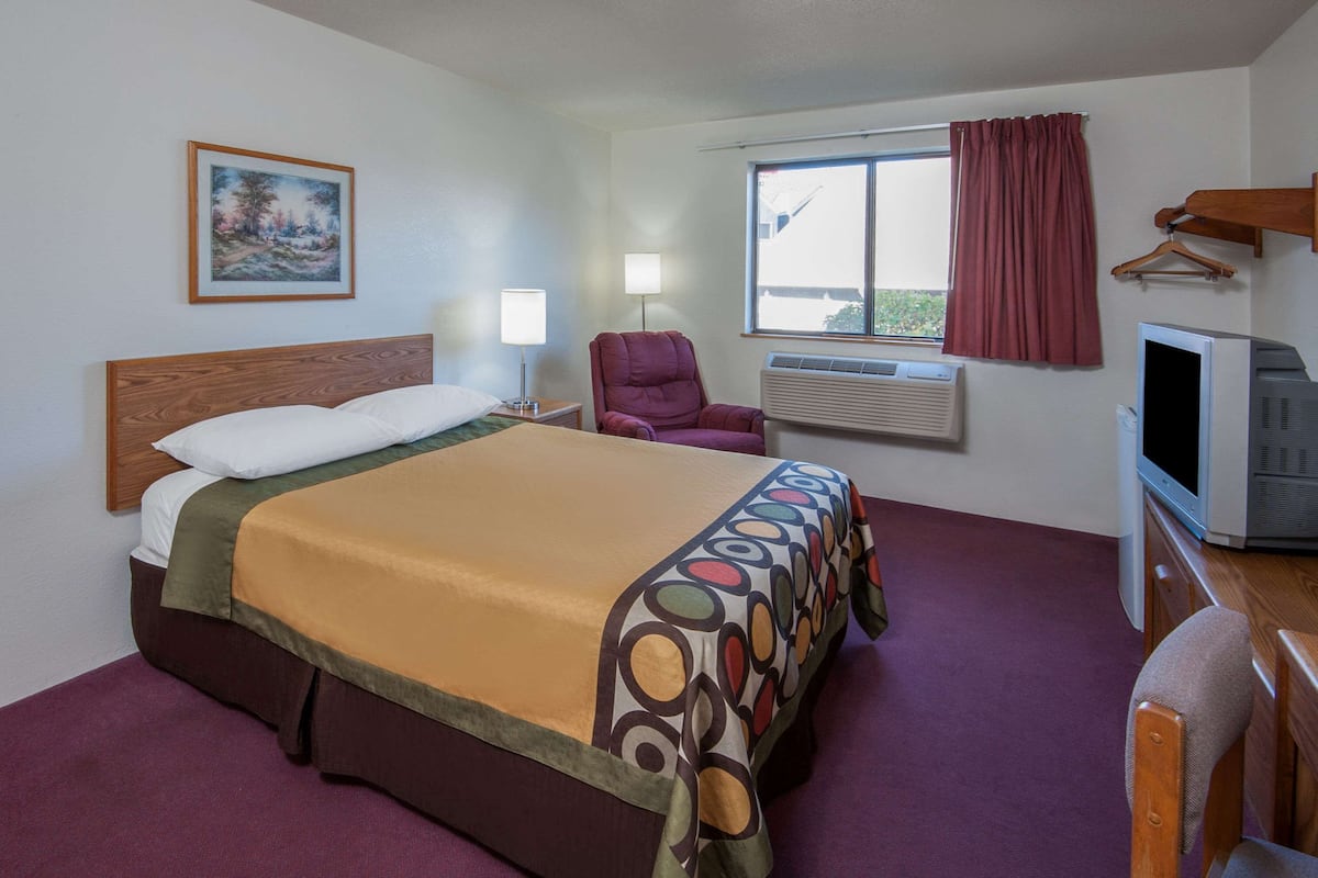 Habitación de hotel con cama, una silla, escritorio con televisor, ventana con cortinas, unidad de aire acondicionado y un cuadro en la pared.