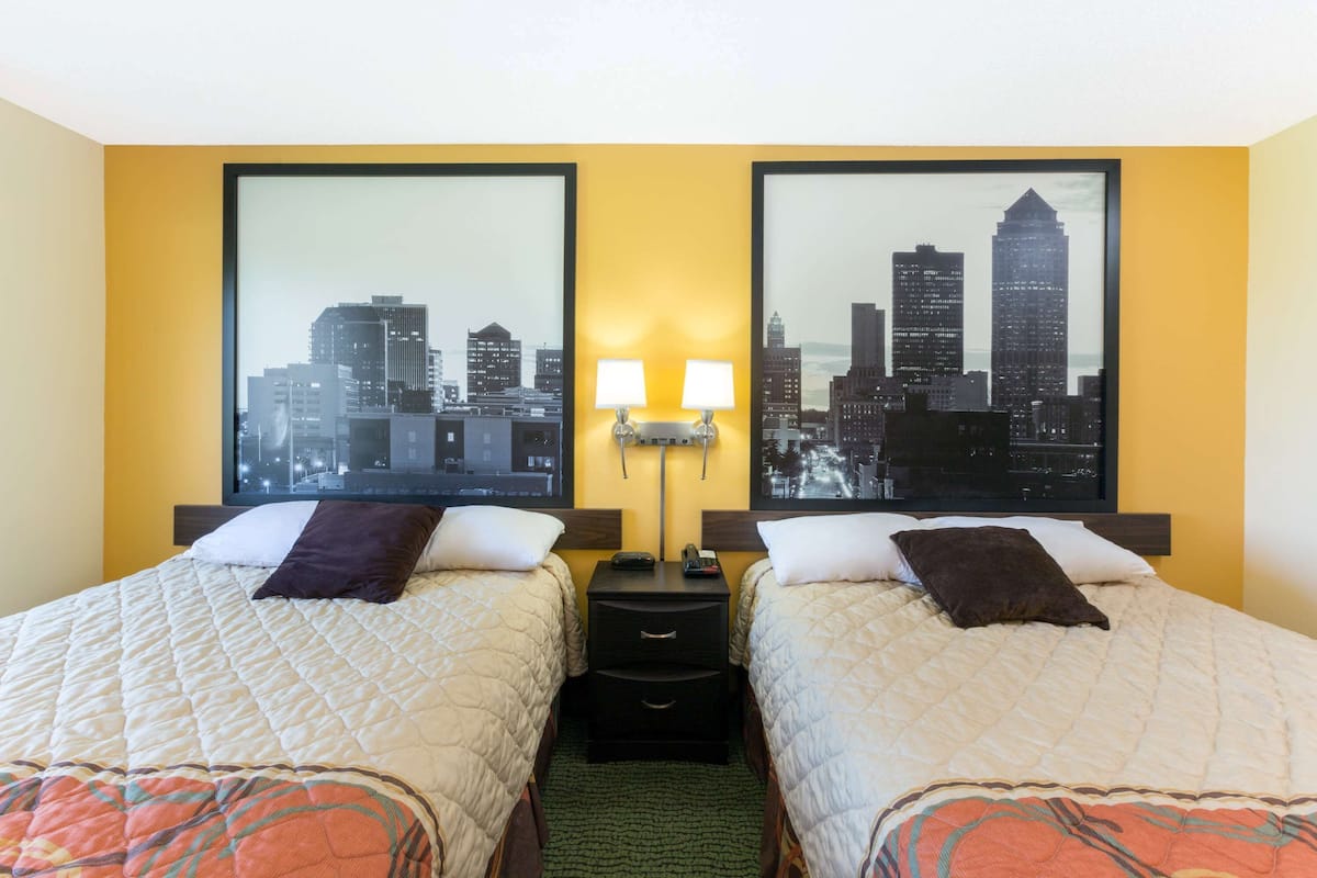 A hotel room with two beds, a nightstand with lamps, and a cityscape view on the wall.