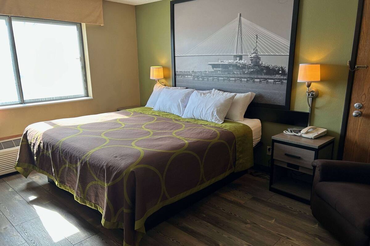 A hotel room with a bed, two bedside lamps, a nightstand with a phone, and a window with a view of a bridge.