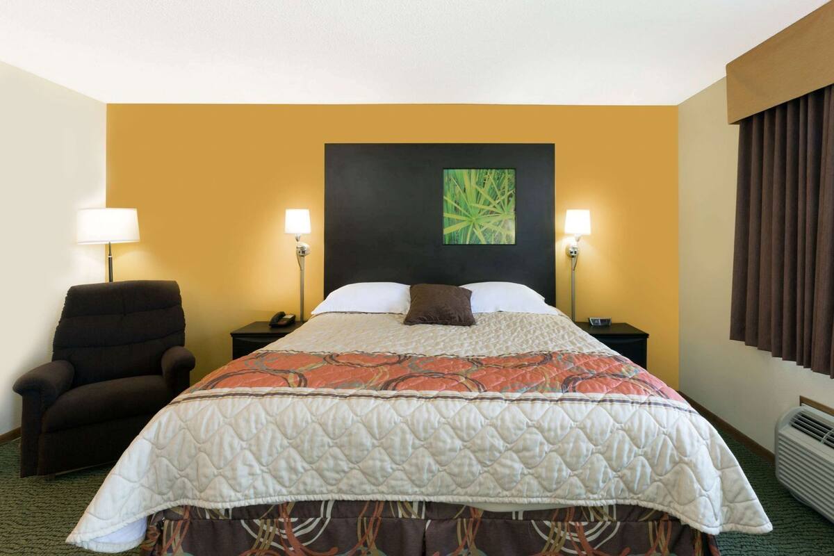 A hotel room with a bed, two bedside lamps, a chair, a wall art piece, and a window with curtains.