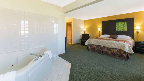 A hotel room with a large bathtub, a bed with a patterned bedspread, two bedside lamps, and a green carpet. - Super 8 by Wyndham Perry IA (Perry)