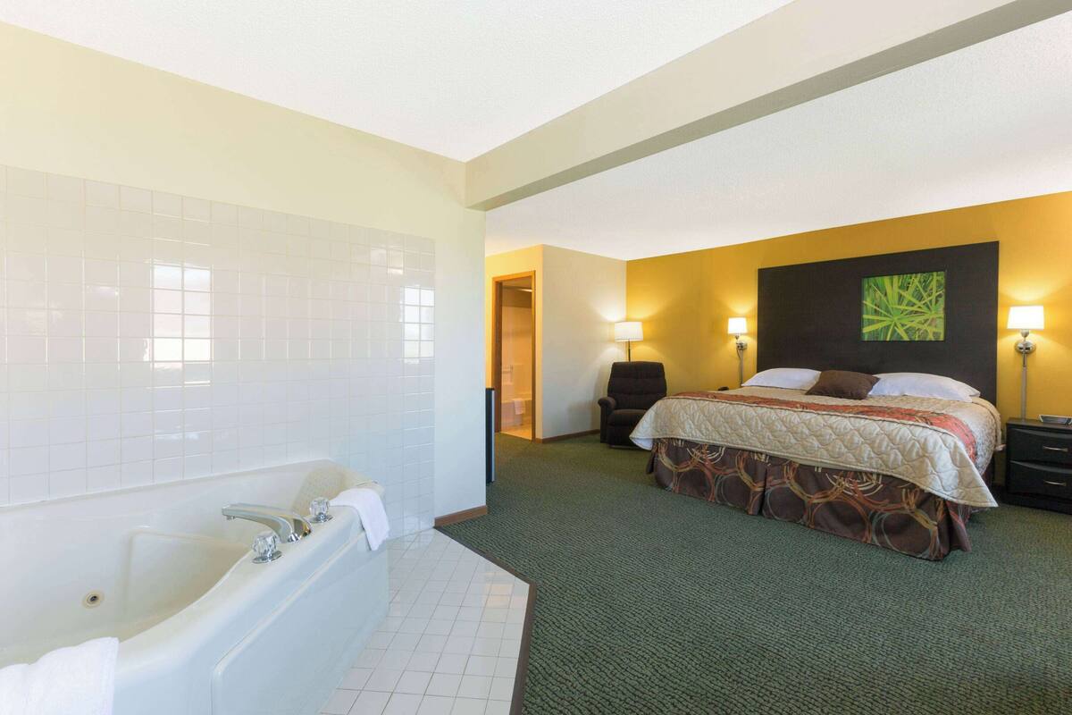 A hotel room with a large bathtub, a bed with a patterned bedspread, two bedside lamps, and a green carpet.