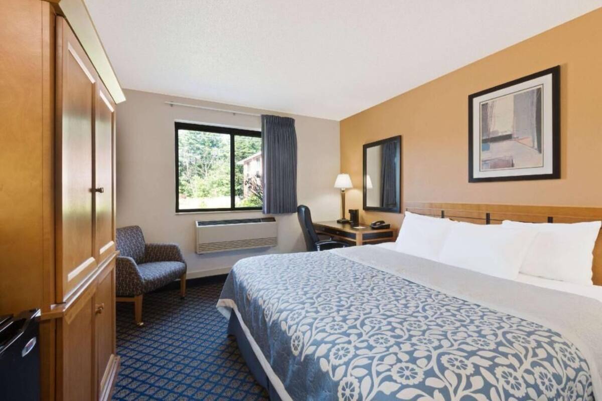 A hotel room with a bed, a desk, a chair, a window with curtains, and a framed picture on the wall.