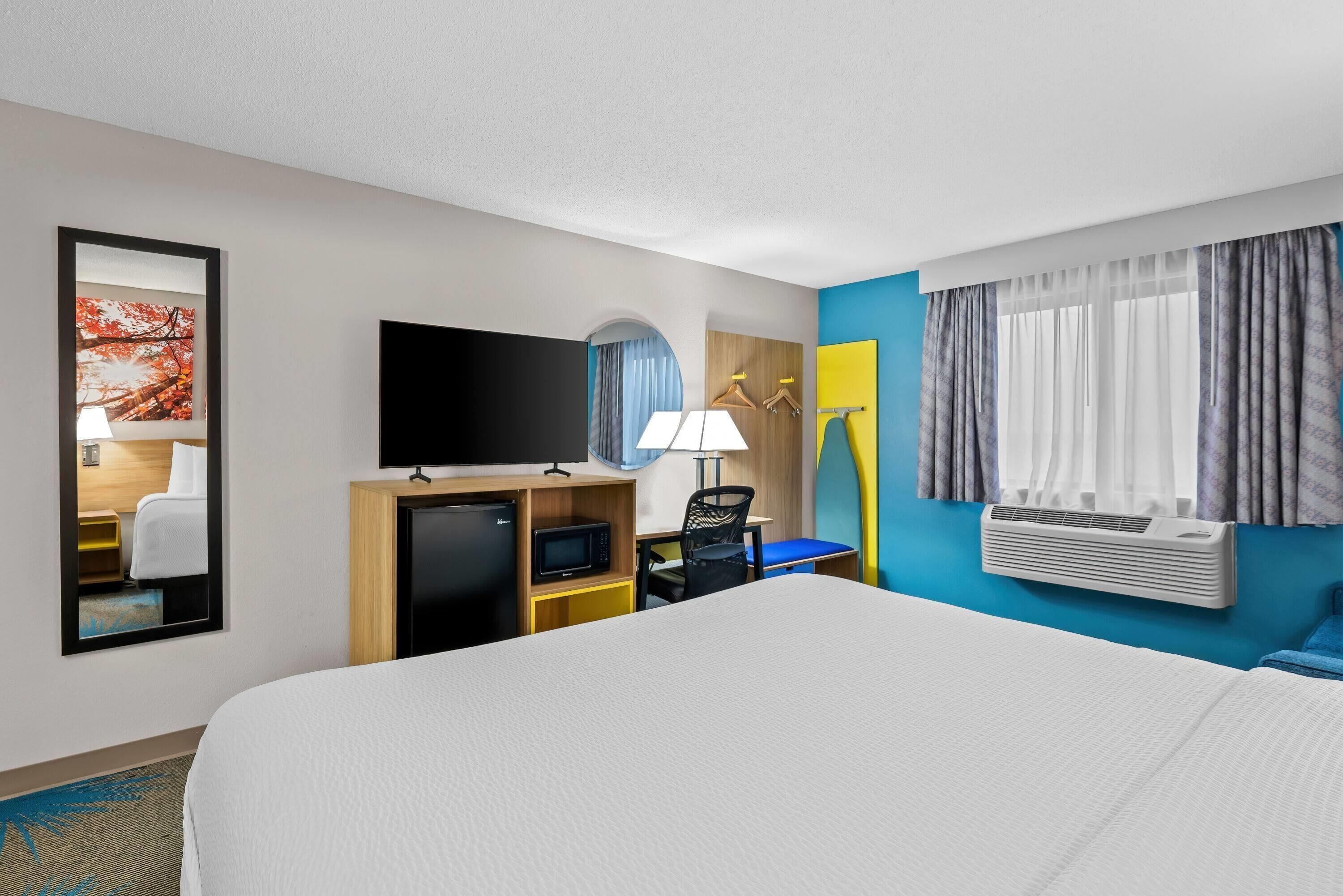 A hotel room with a bed, a desk with a TV, a mini-fridge, a lamp, and a window with curtains.