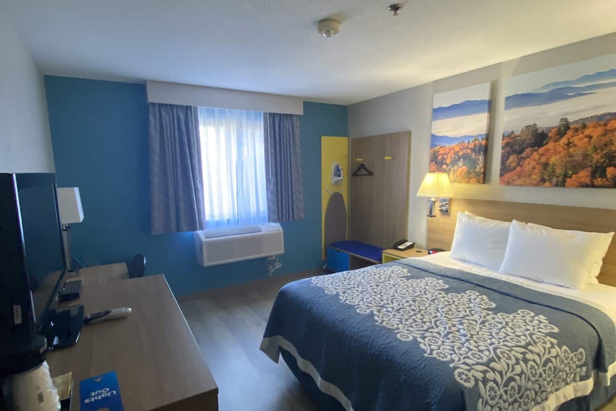 A hotel room with a bed, a desk, a chair, a window with curtains, a painting on the wall, and a door.