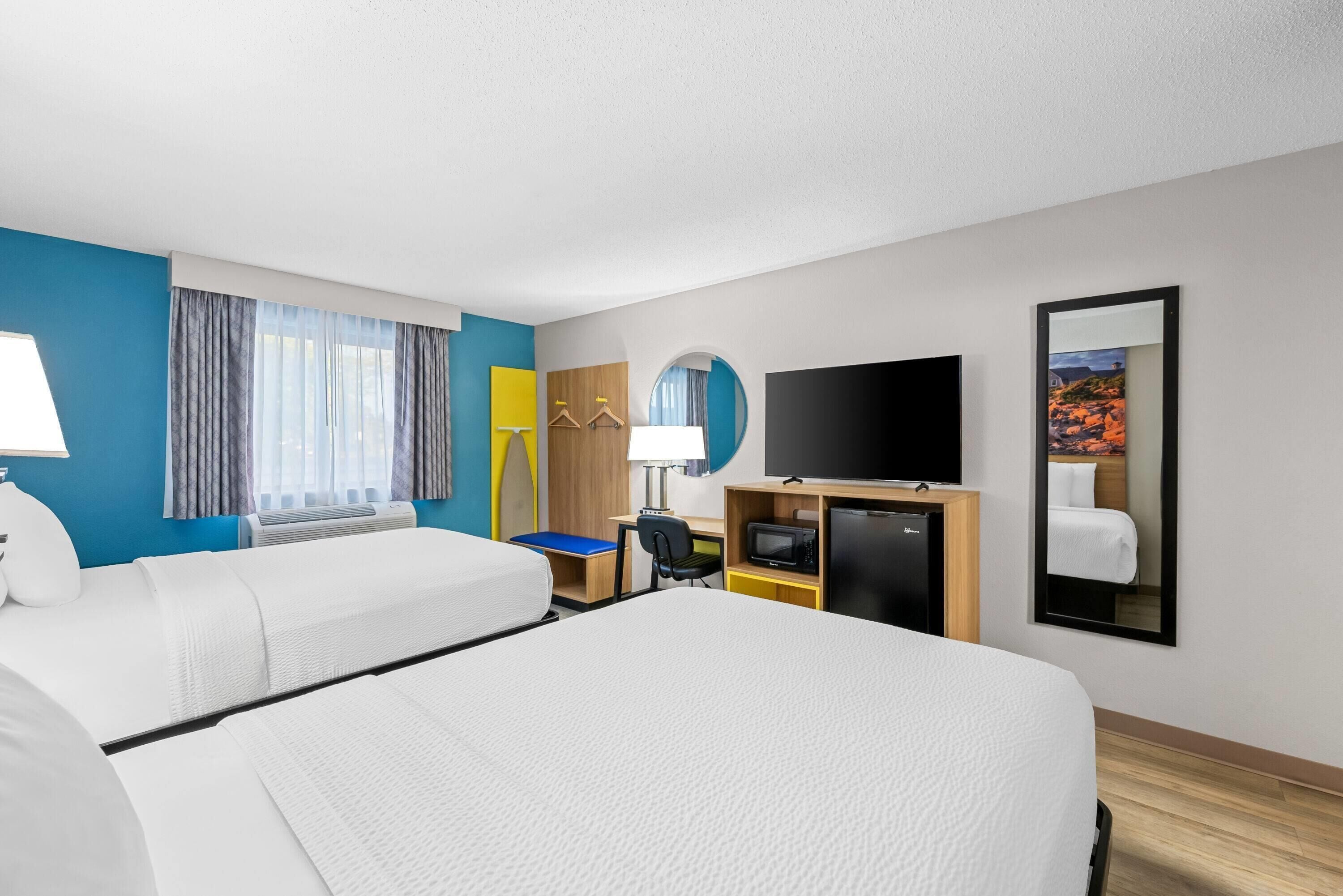 A hotel room with two beds, a desk, a TV, a mirror, and a window with curtains.