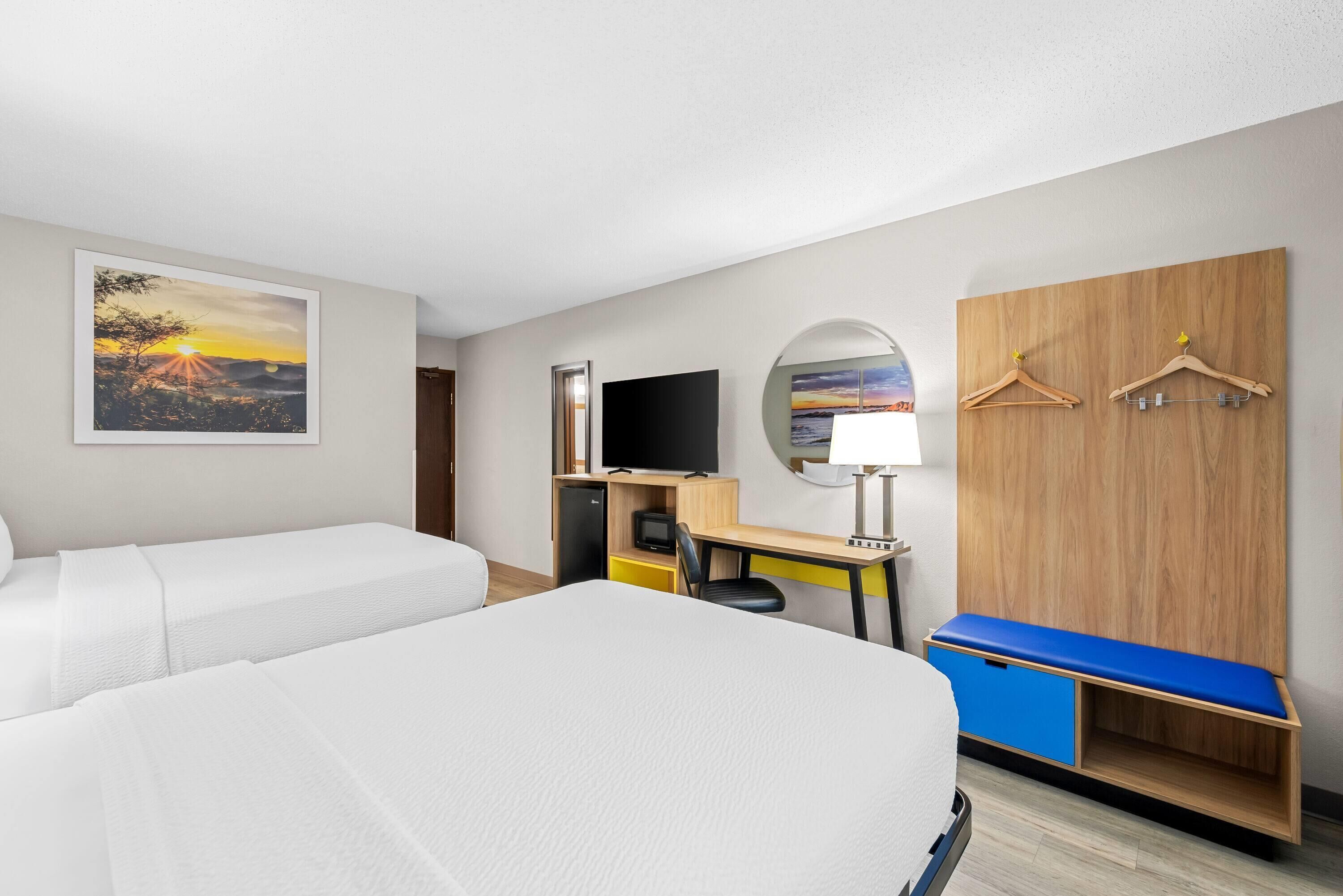 A hotel room with two beds, a desk with a chair, a TV, a wardrobe, and a painting of a sunset.