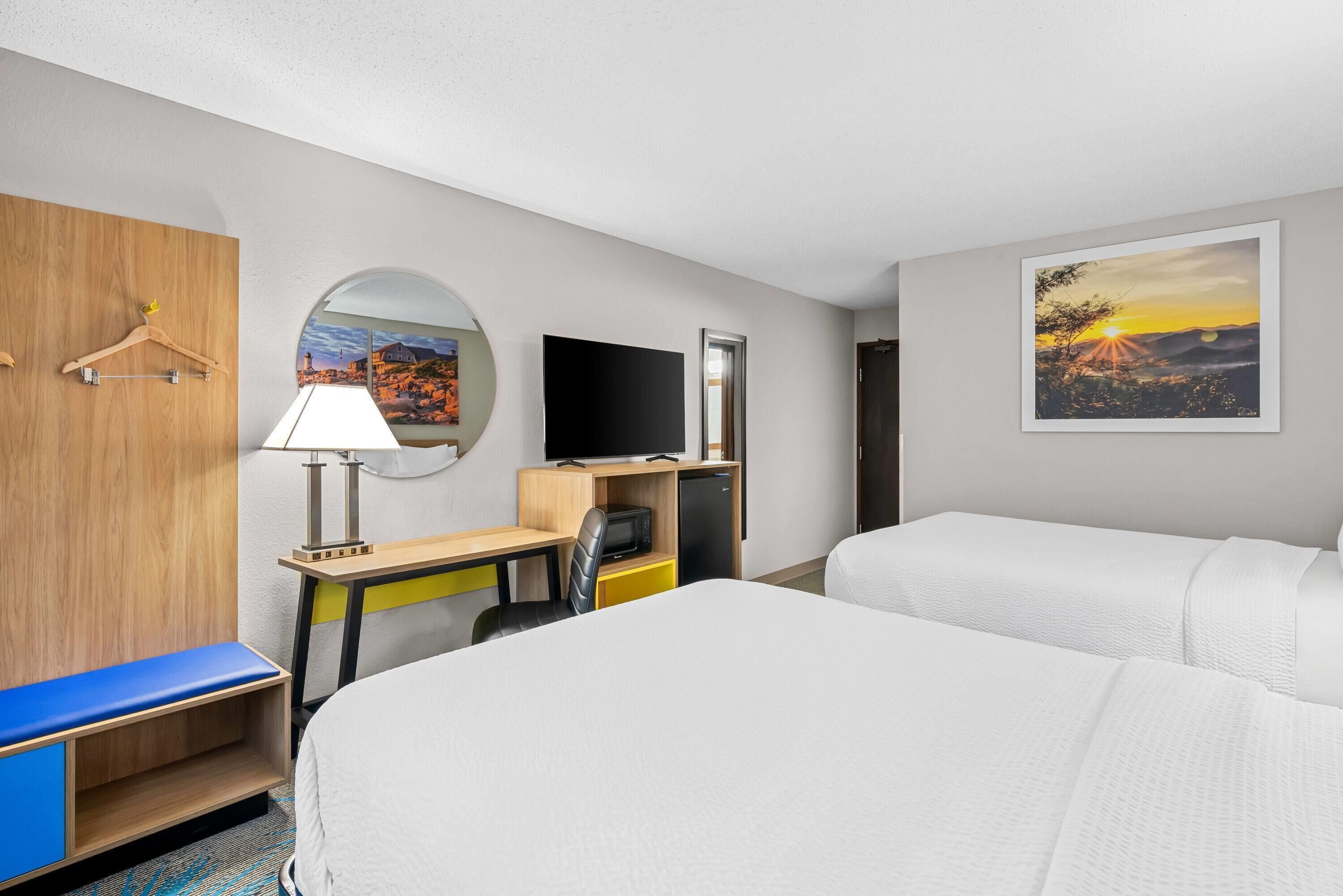 A hotel room with two beds, a desk with a chair, a TV, and a painting of a sunset.