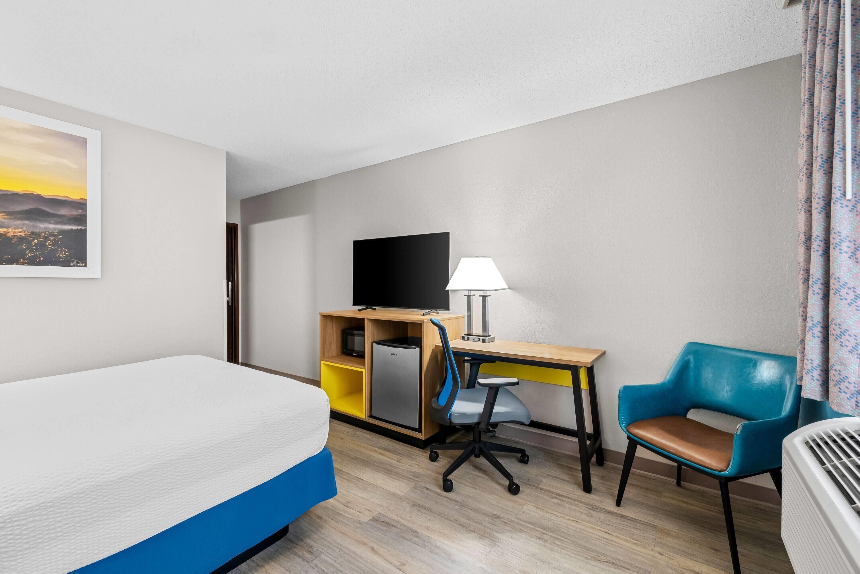 A hotel room with a bed, desk, chair, TV, and a small kitchenette.